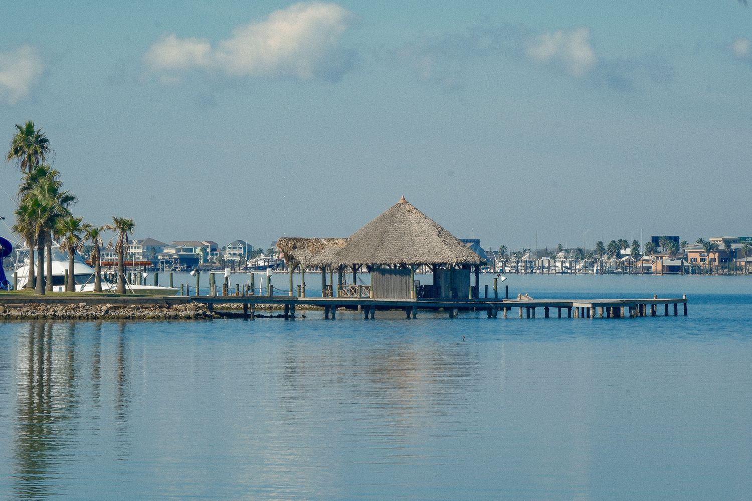 Thatched roof tiki hut on a wooden pier over calm blue water with palm trees and waterfront homes.