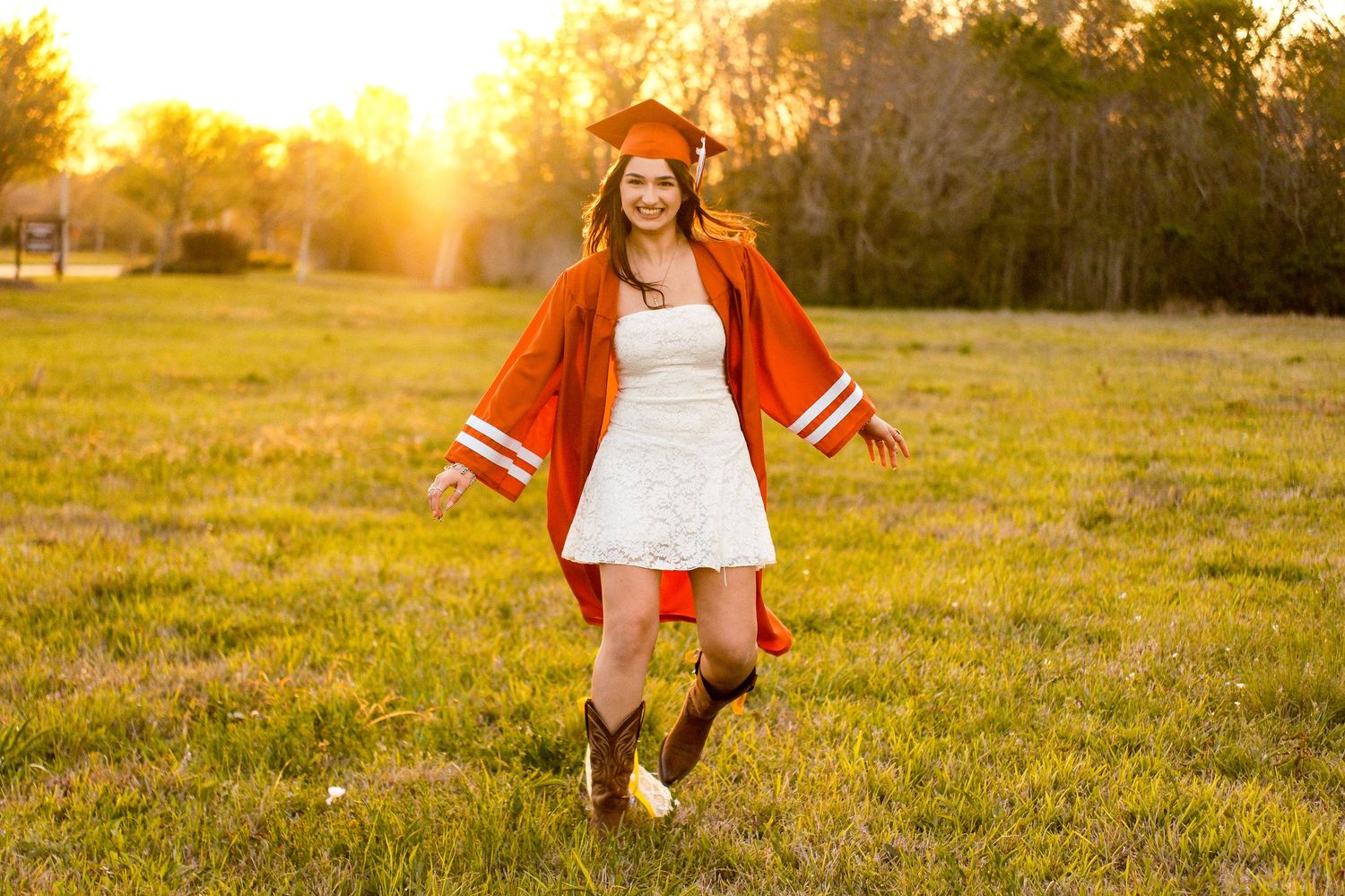 Graduate in orange cap and gown over white dress and cowboy boots celebrates outdoors at golden hour.