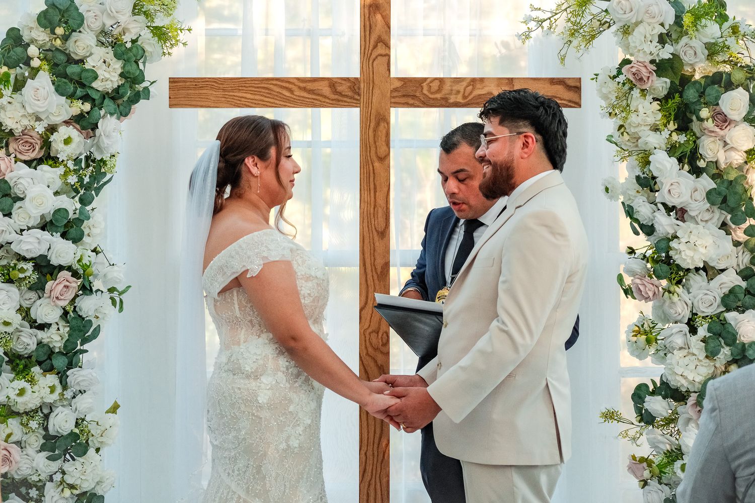 Bride and groom hold hands at wedding altar adorned with white flowers and wooden cross during ceremony.