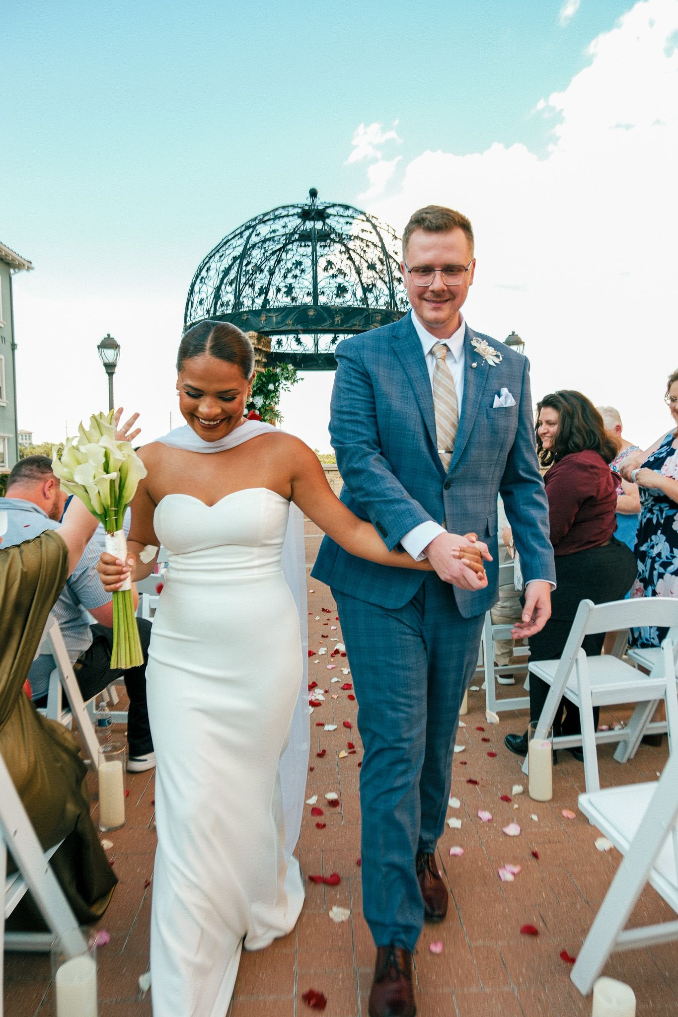 Joyful newlywed couple walking down outdoor aisle after wedding ceremony with rose petals on ground.
