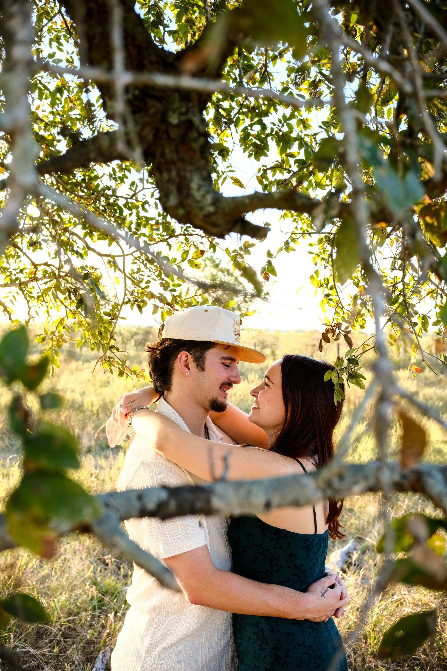 A couple embraces and smiles at each other under a sunlit tree in a golden field.