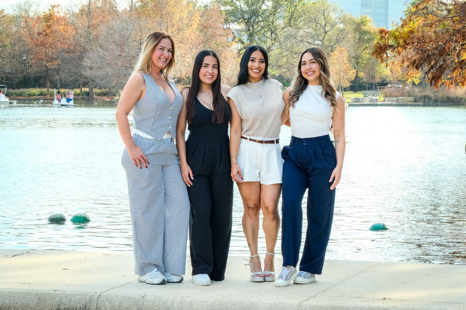 Four stylish women posing together by a lake with autumn trees in the background on a sunny day.