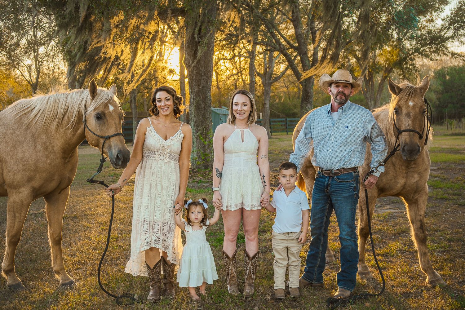 Family portrait with two palomino horses at golden sunset in a wooded outdoor setting.