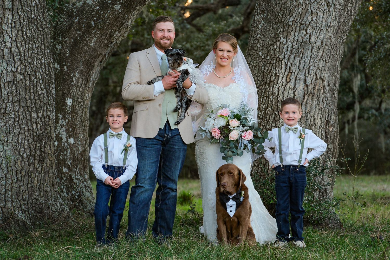 Wedding family portrait with groom, bride, two young boys in suspenders, and dogs under large oak trees.