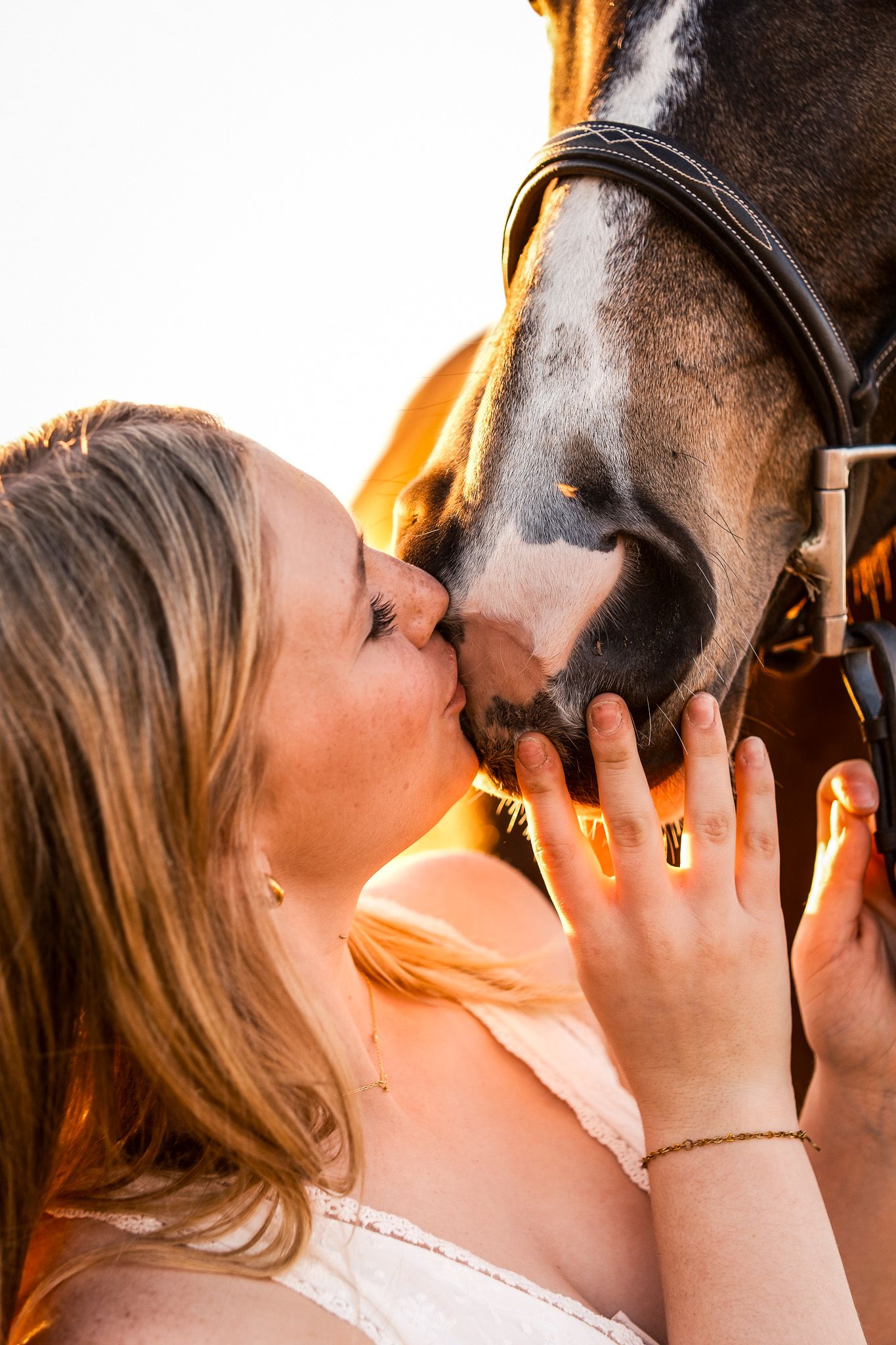 Young blonde woman gently kissing a dark horse on the nose in a warm golden backlit setting.