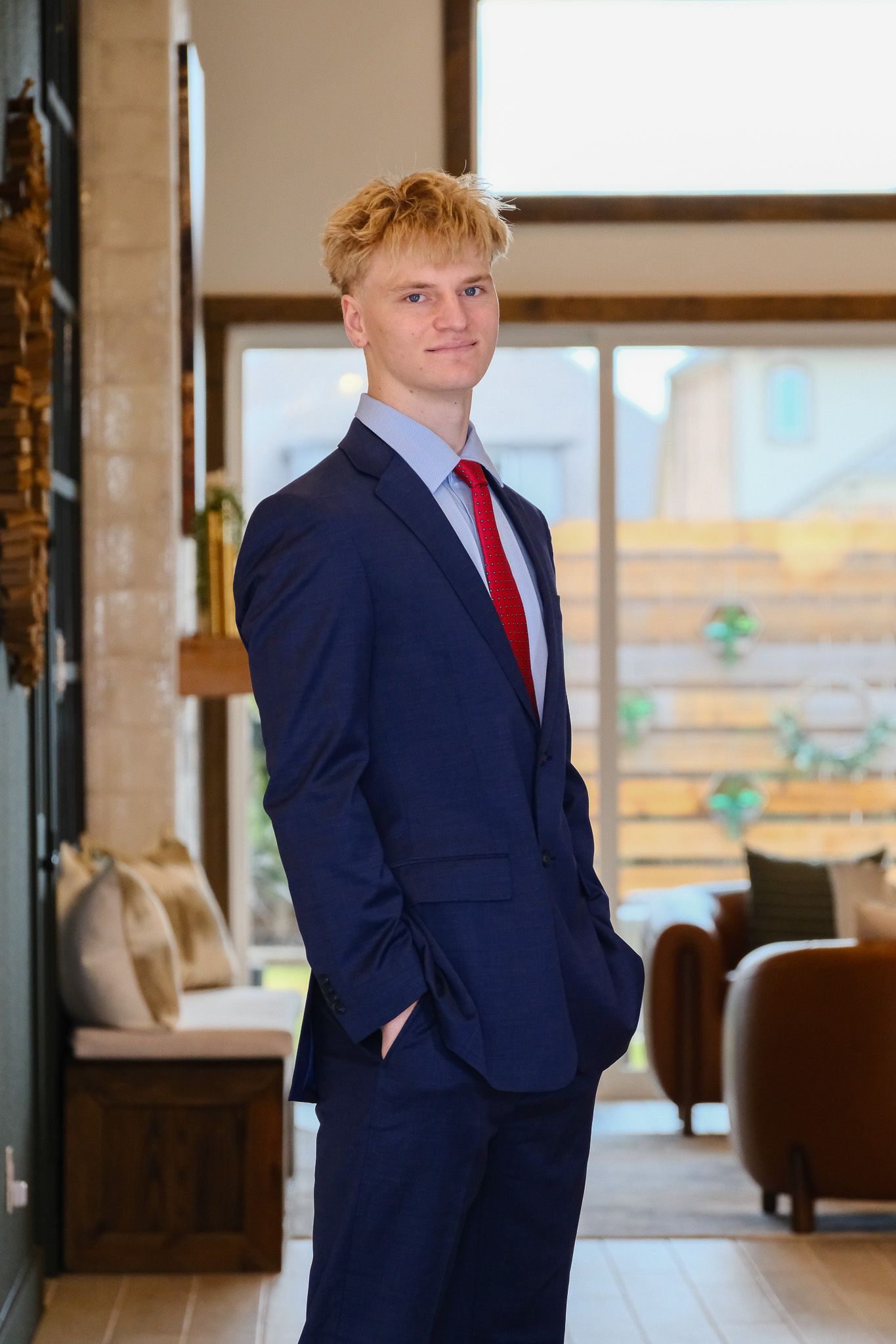 Young man in navy blue suit and red tie posing indoors with hands in pockets.