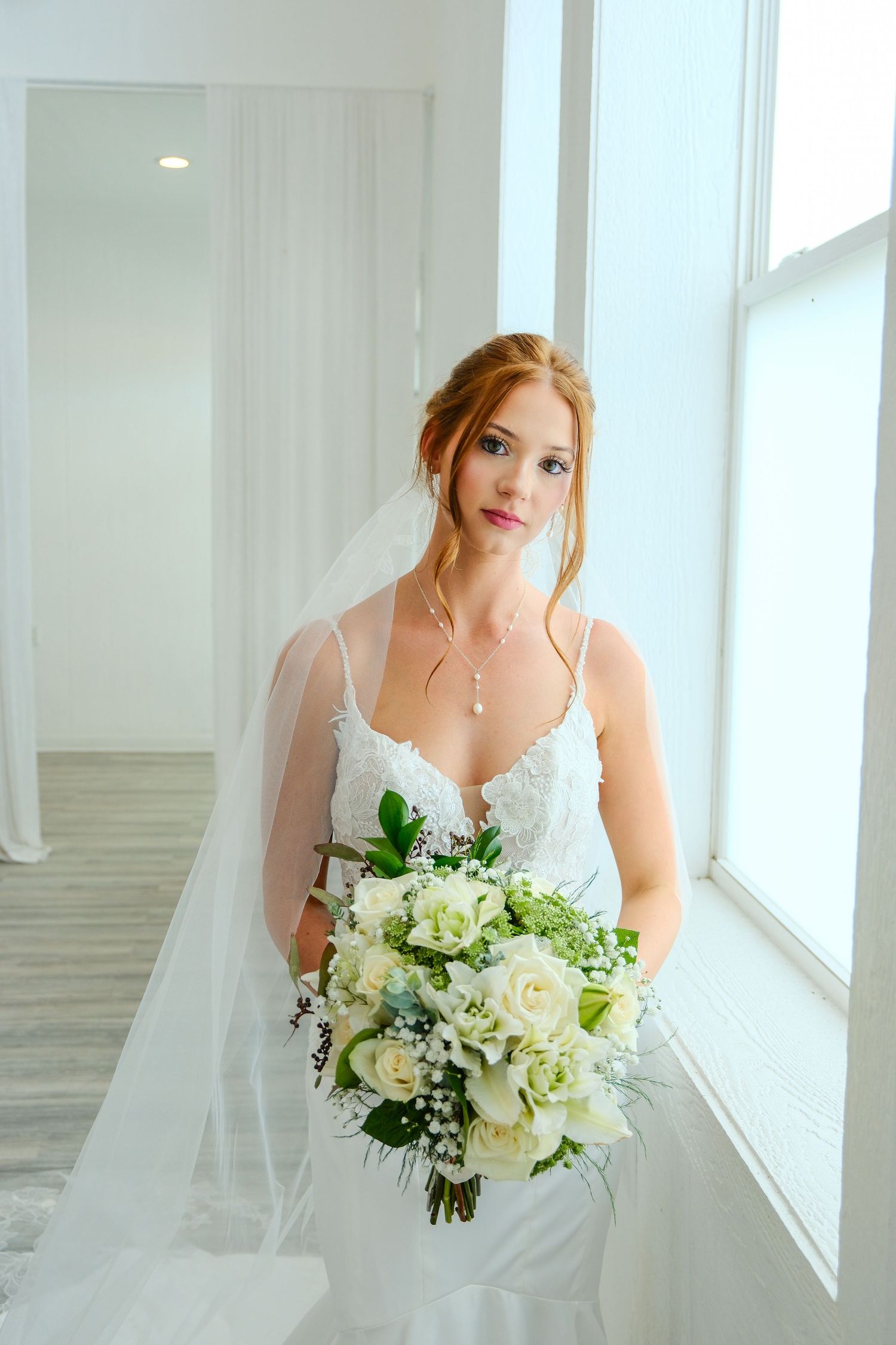 Bride in white lace gown holding bouquet of white roses and greenery, standing by bright window with veil.