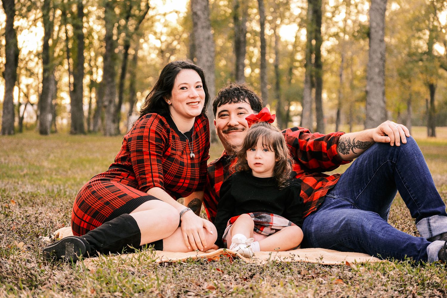Happy family of three in matching red plaid shirts relaxing on grass in autumn park.