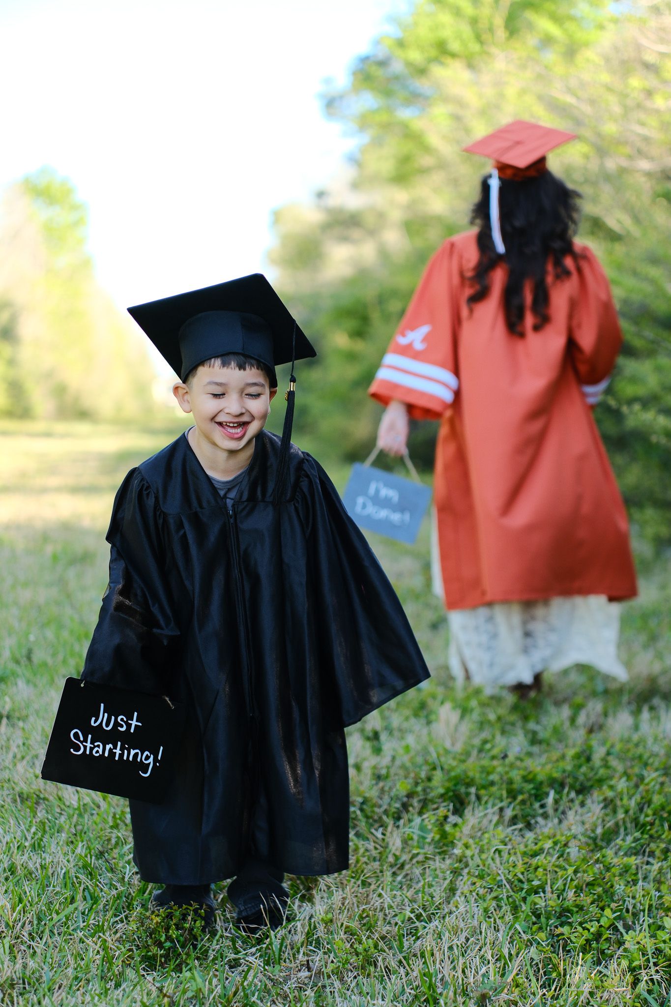 Two children in graduation caps and gowns, one holding a Just Starting sign, celebrate outdoors.