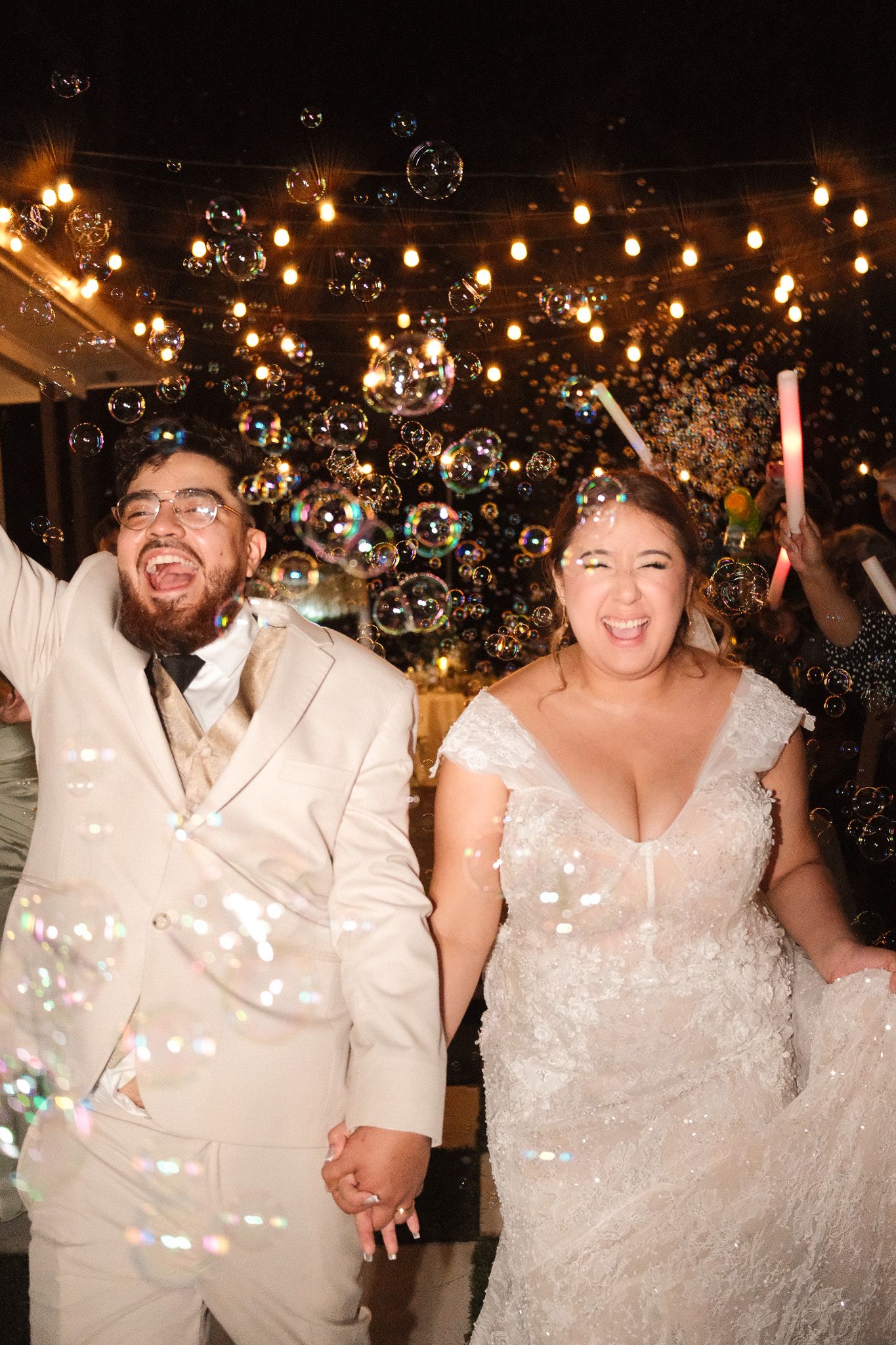 Joyful bride and groom walk through bubbles holding hands during festive outdoor wedding celebration at night.