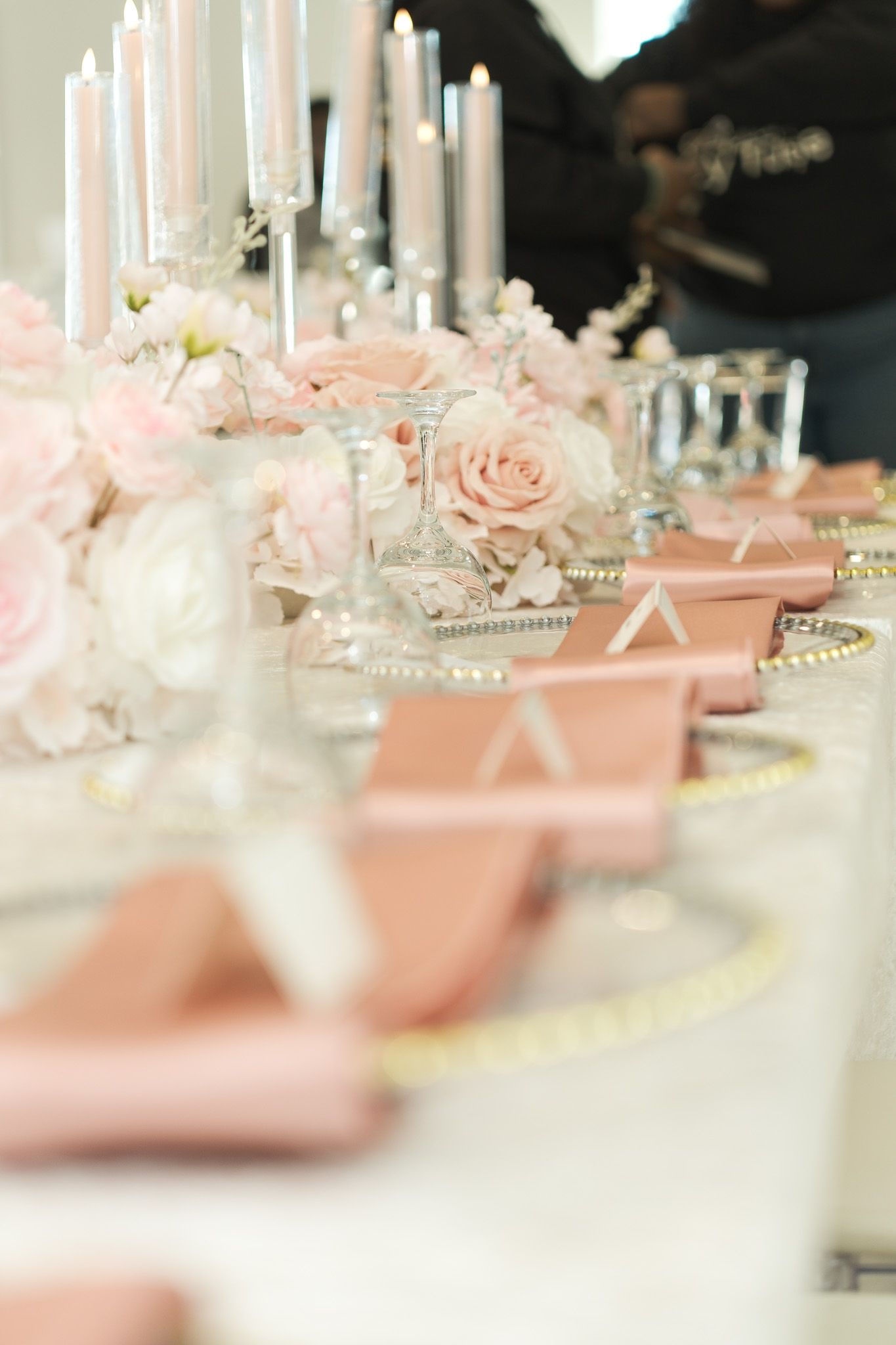 Elegant wedding reception table with pink roses, white candles, gold charger plates, and blush pink napkins.