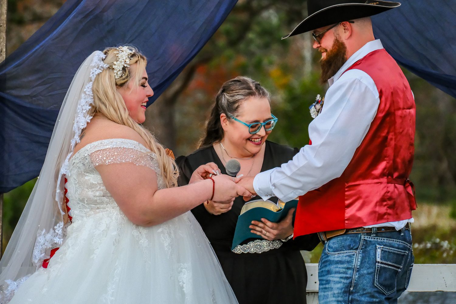 Groom in red vest and cowboy hat kissing bride under decorated arch with string lights.