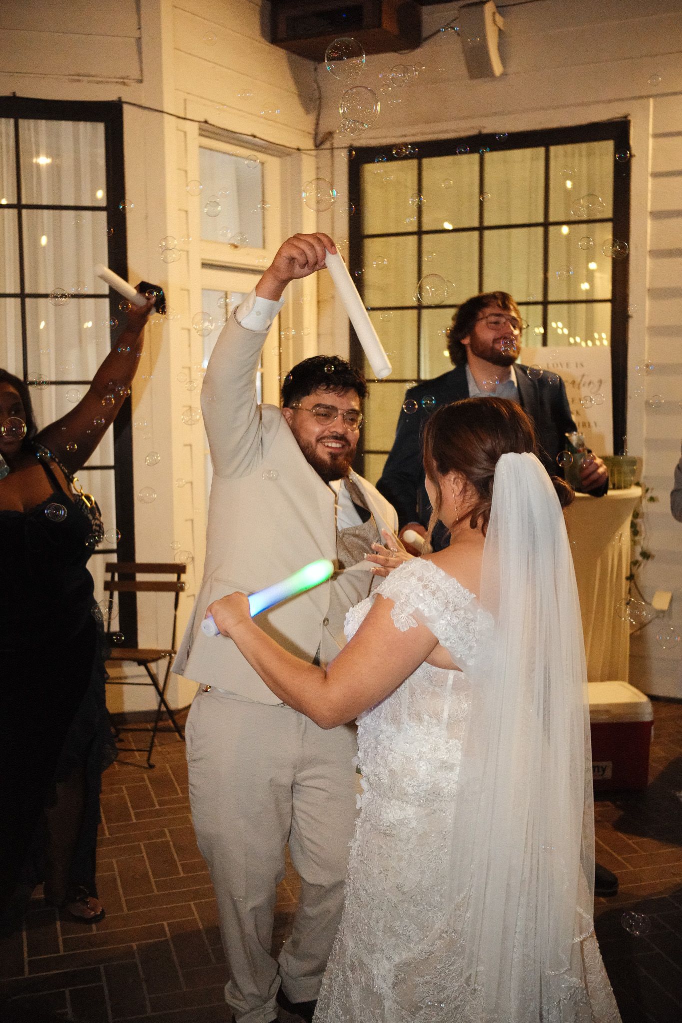 Groom in tan suit dances with bride surrounded by bubbles and guests at lively wedding reception.