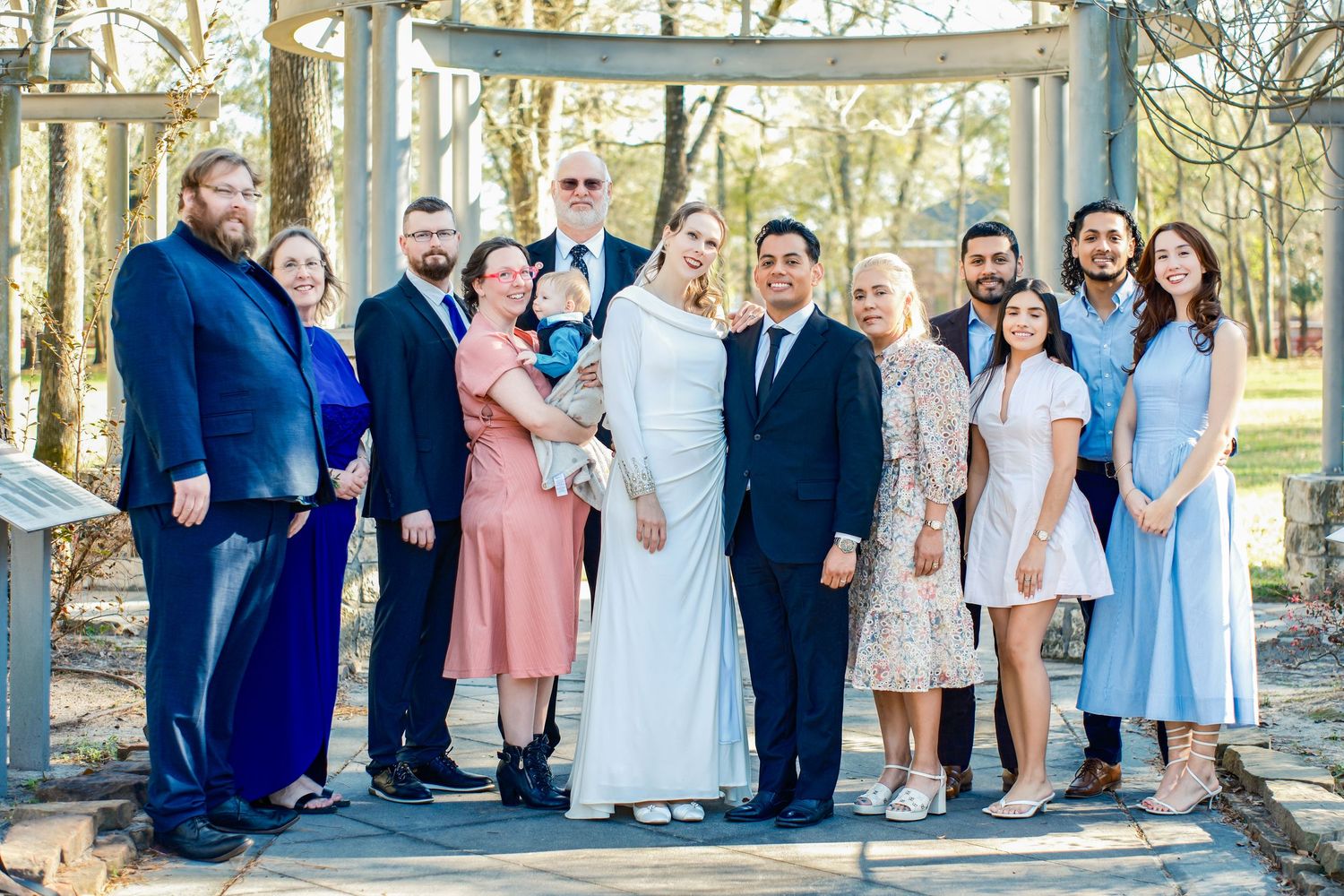 A joyful wedding party poses outdoors under a wooden arbor, featuring the bride in white and groom surrounded by family.