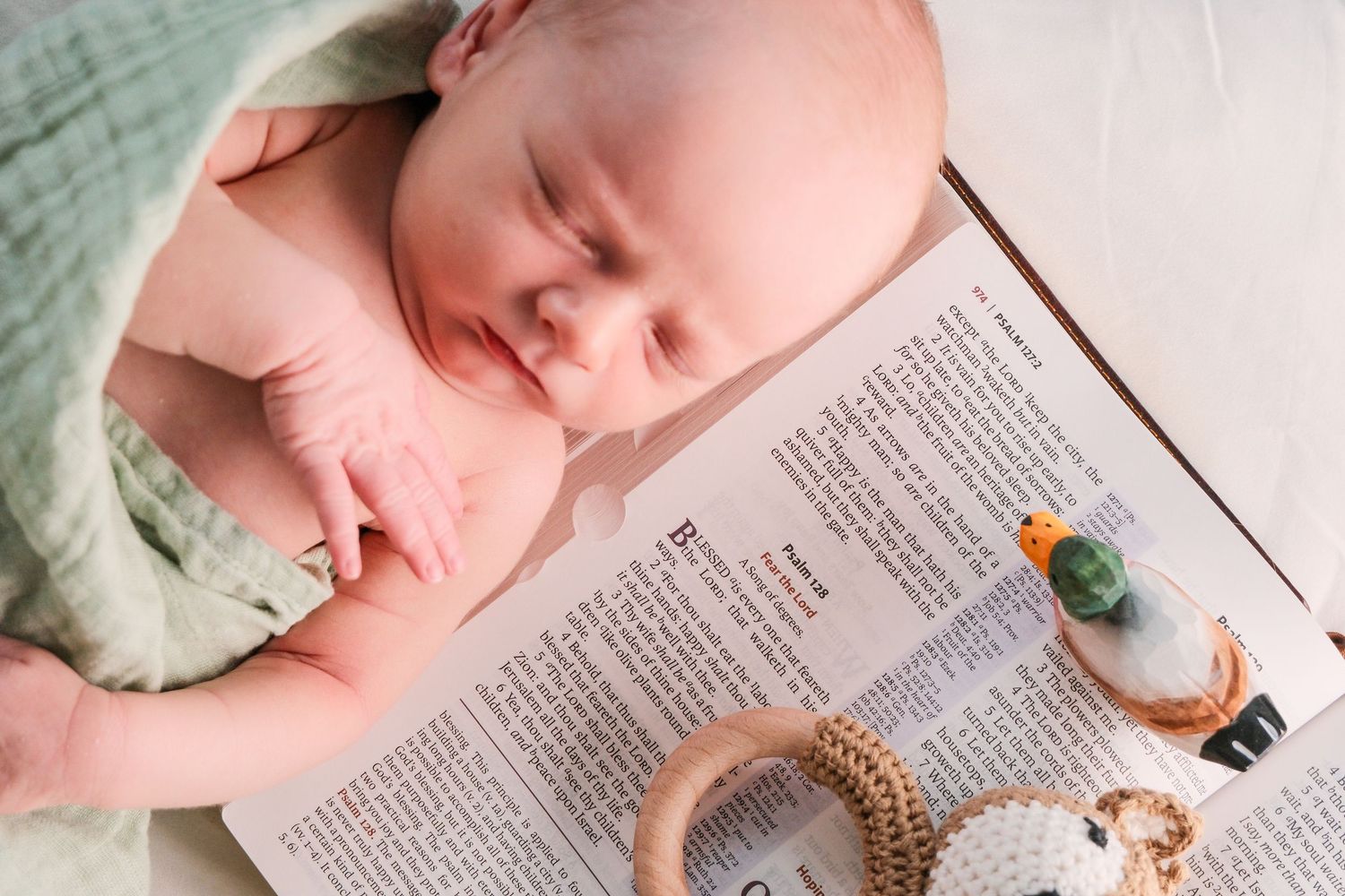 Sleeping newborn baby resting on an open Bible with a wooden teething ring and small toy nearby.