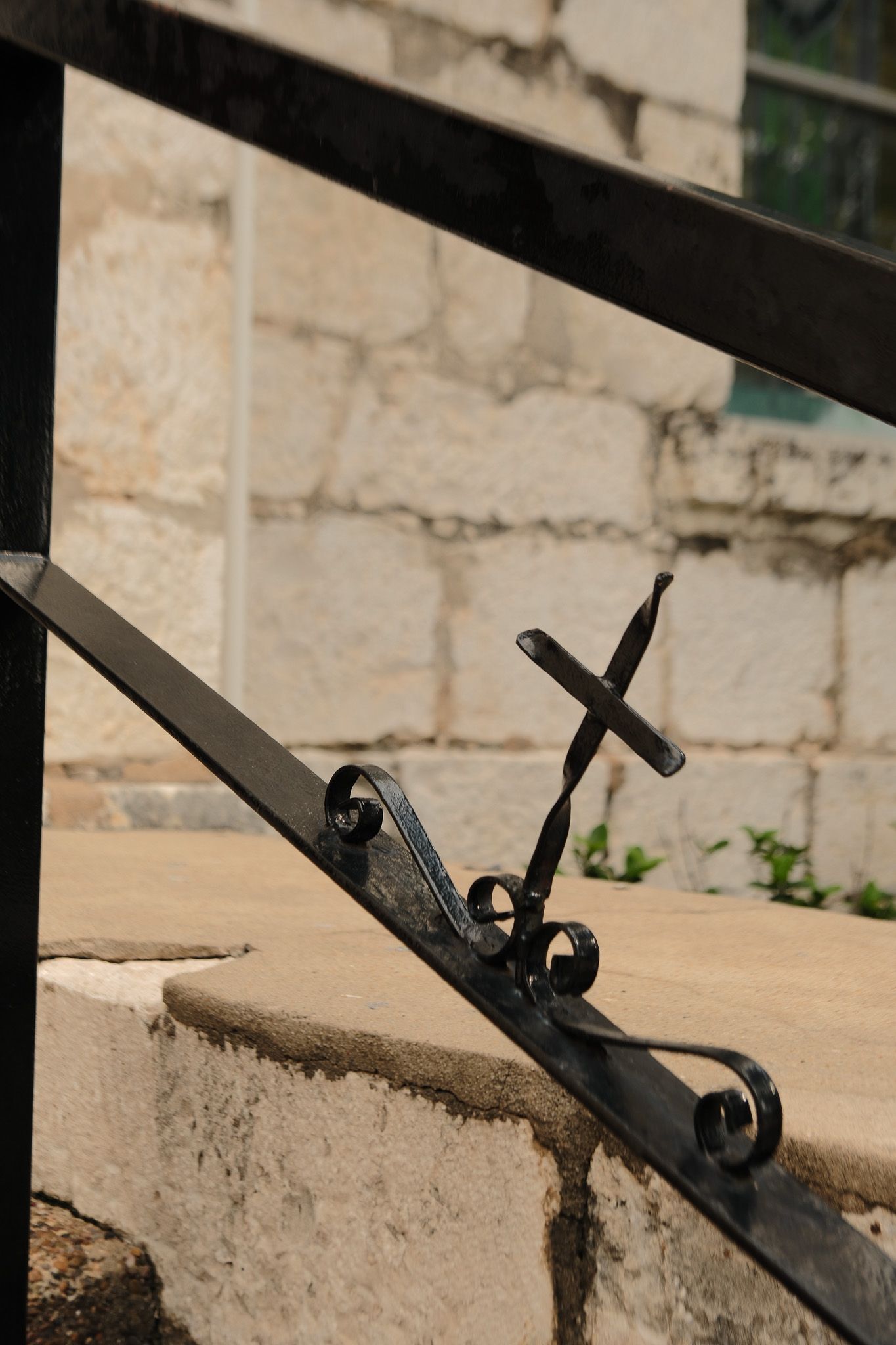 Ornate black wrought iron cross detail on a railing with stone wall background in sunlight.