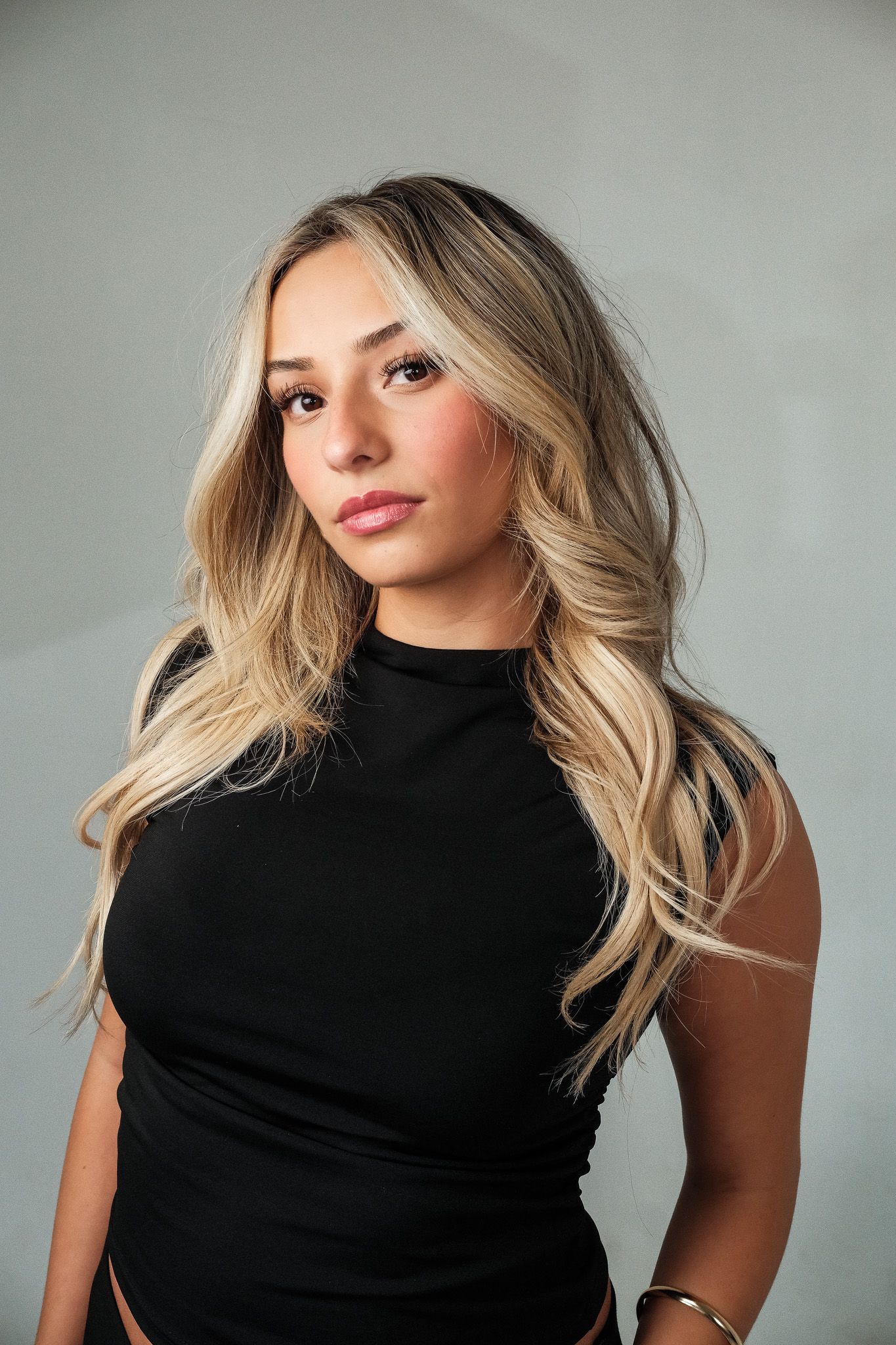 Young blonde woman in black sleeveless turtleneck poses against gray studio background.