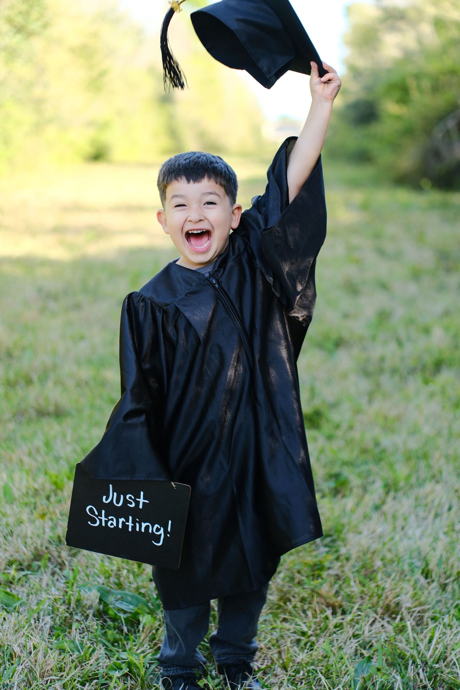 Excited young boy in graduation gown holds cap up holding Just Starting chalkboard sign.