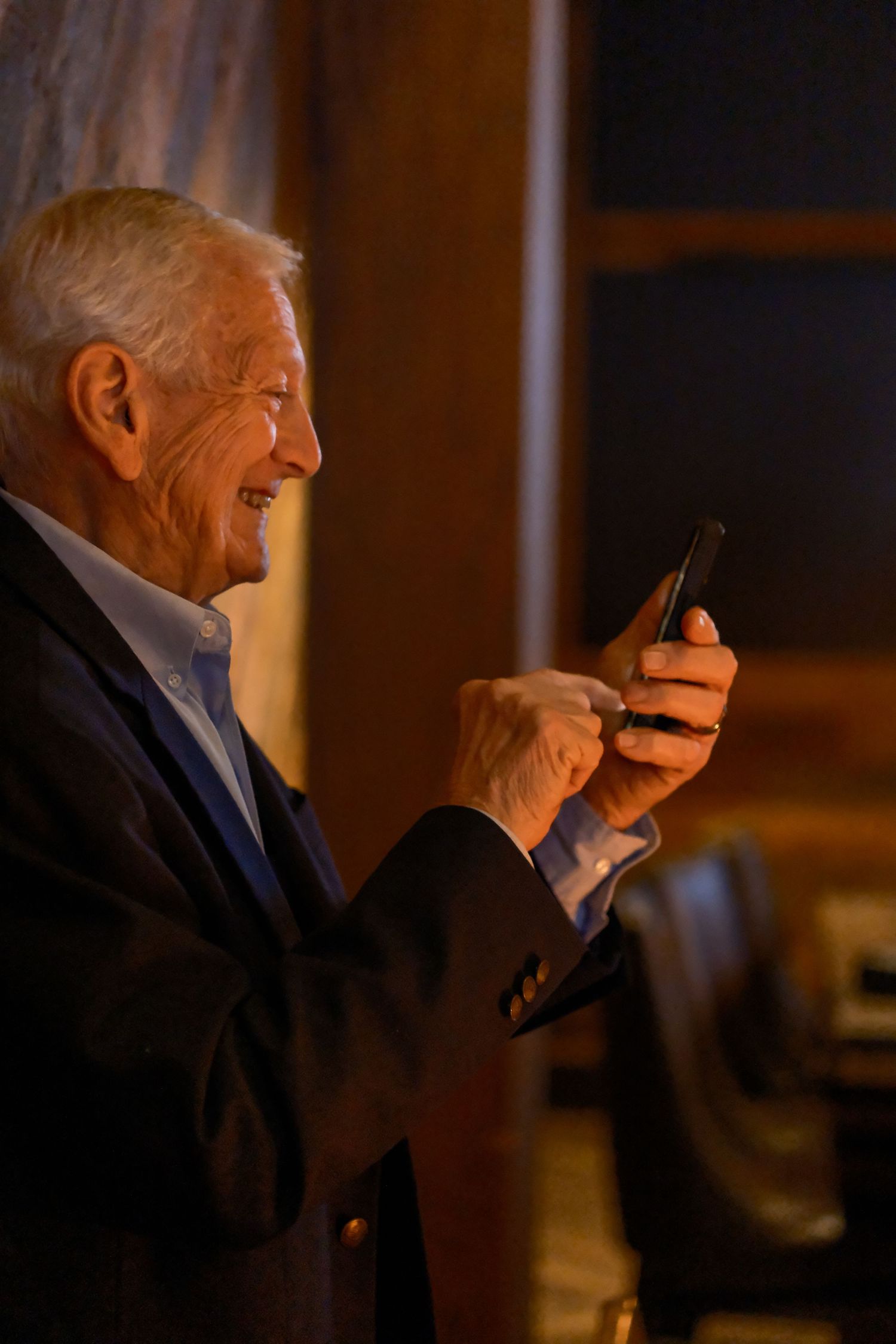 Smiling elderly man in dark suit using smartphone indoors with warm ambient lighting.