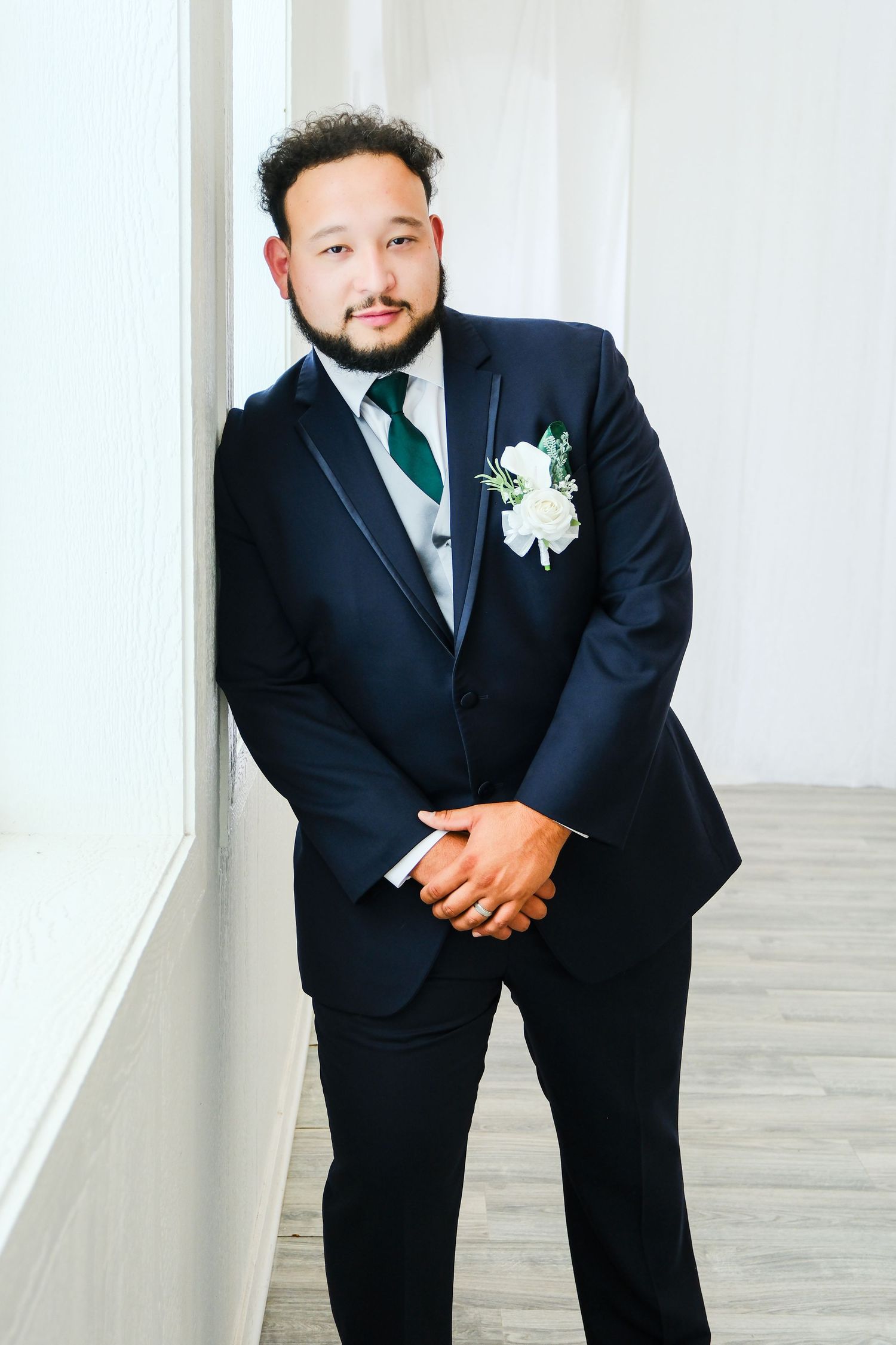 A well-dressed groom in a navy suit with green tie and white boutonniere leans against a white wall, smiling confidently.