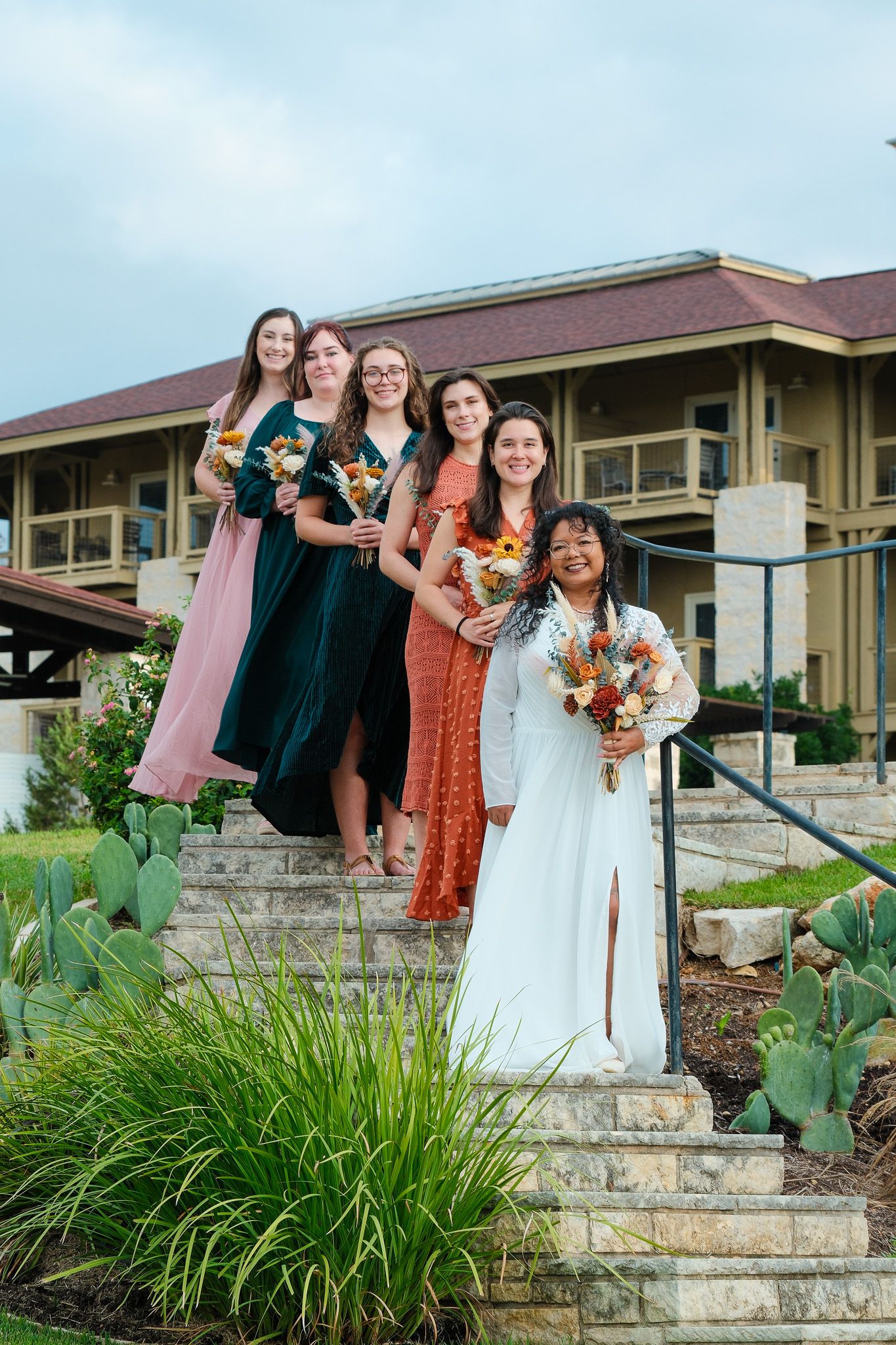 Bride in white gown poses with bridesmaids holding colorful bouquets on stone steps at outdoor venue.