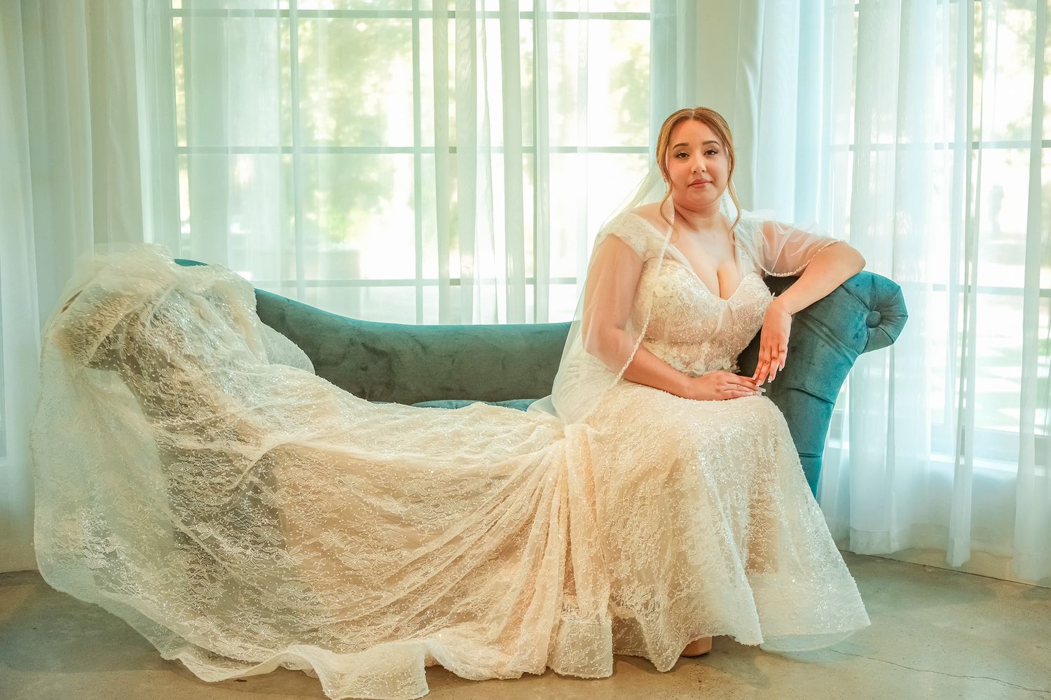 Elegant plus-size bride in lace wedding gown sitting gracefully on a teal velvet chaise lounge.