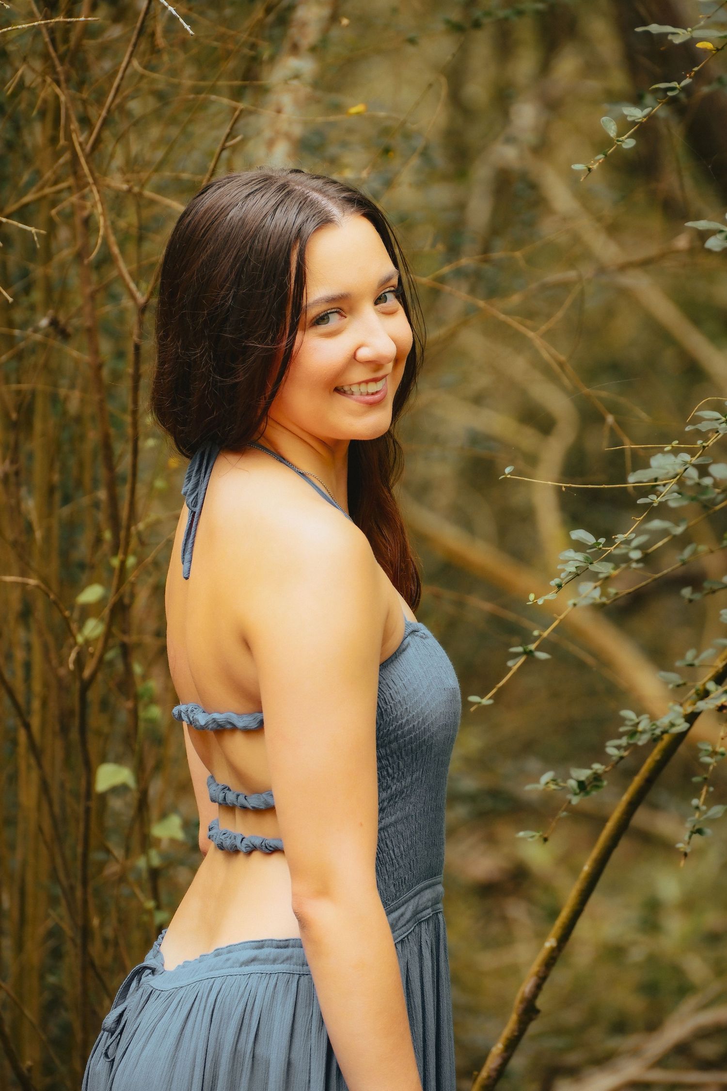 Smiling brunette woman in a blue halter dress poses outdoors among trees and foliage in a natural forest setting.