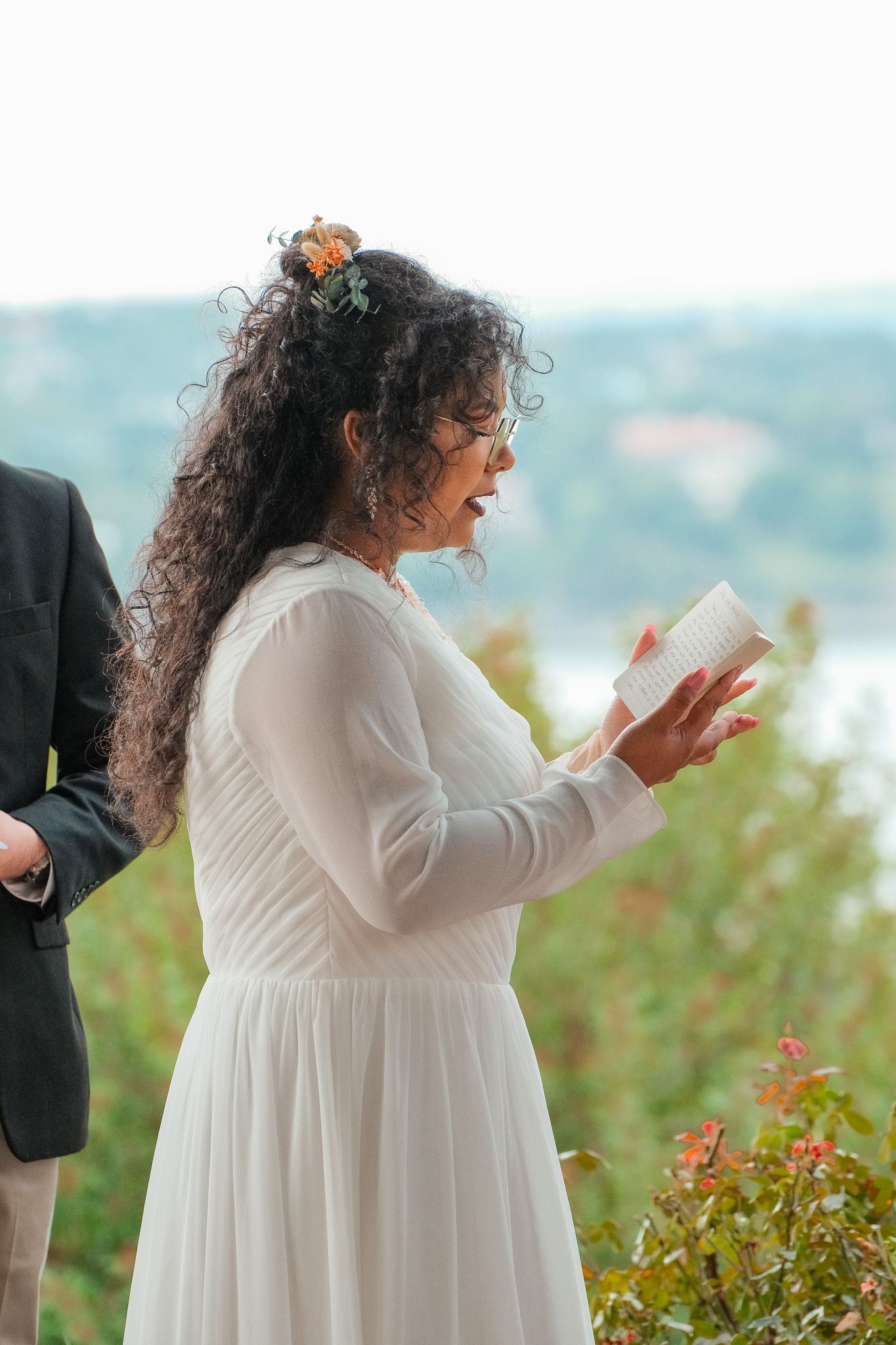 A bride in a white dress reads her wedding vows from a small booklet with a lake visible in the background.