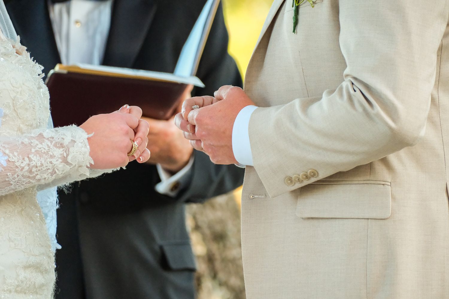Bride and groom exchanging wedding rings during outdoor ceremony with officiant holding book in background.