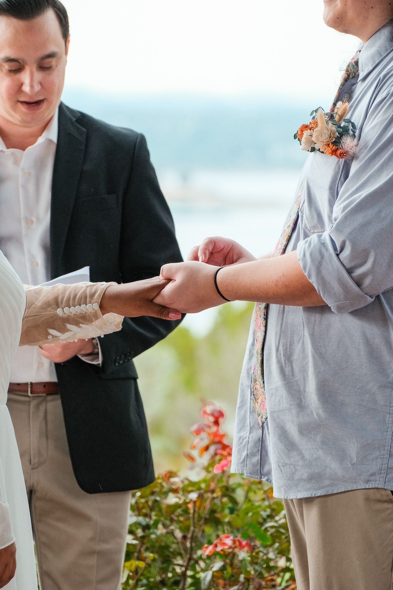 An officiant watches as a groom places a wedding ring on his partners hand during an outdoor ceremony.
