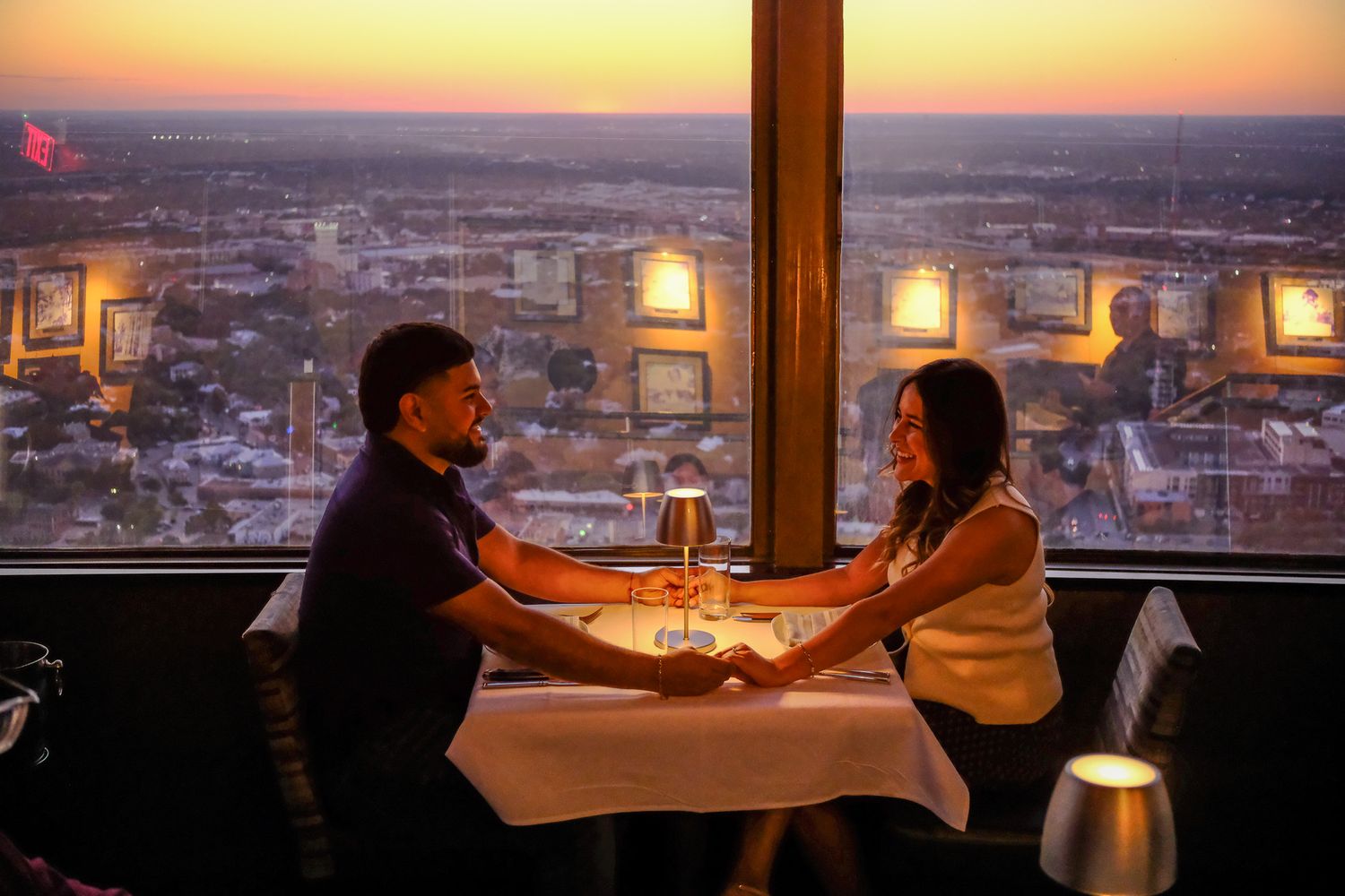 Romantic couple holding hands at a candlelit dinner table with a stunning city sunset view.