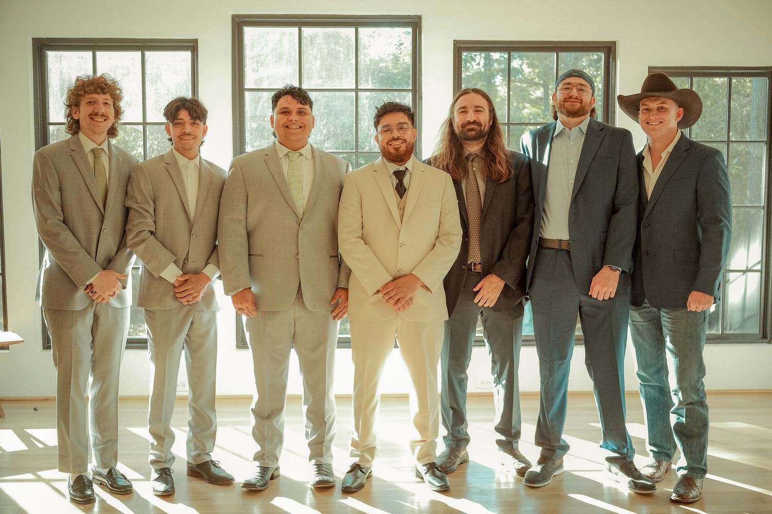 Seven groomsmen posing together in tan and gray suits in a bright sunlit wedding venue.
