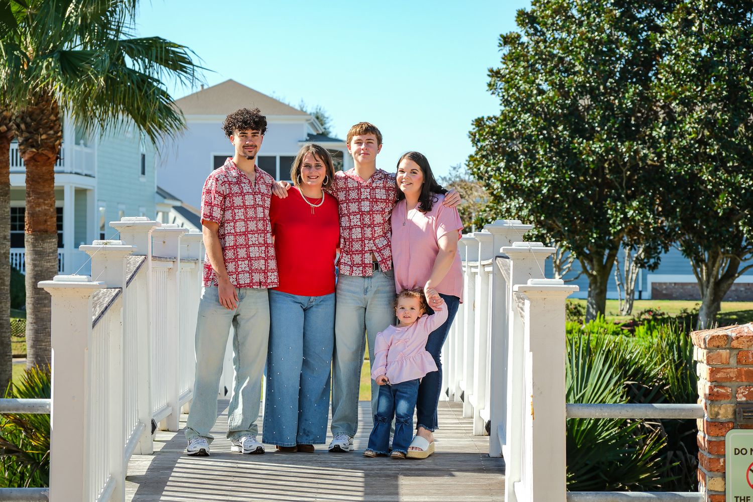 Family of five smiling together on a white wooden bridge outside a coastal home on a sunny day.