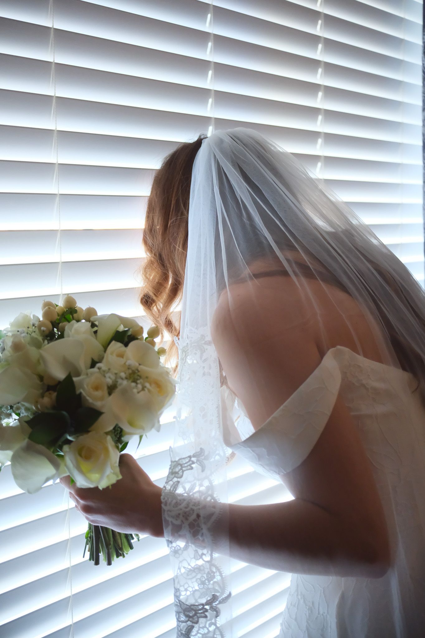 Beautiful bride in white veil holding bouquet of white flowers while standing by window with blinds.