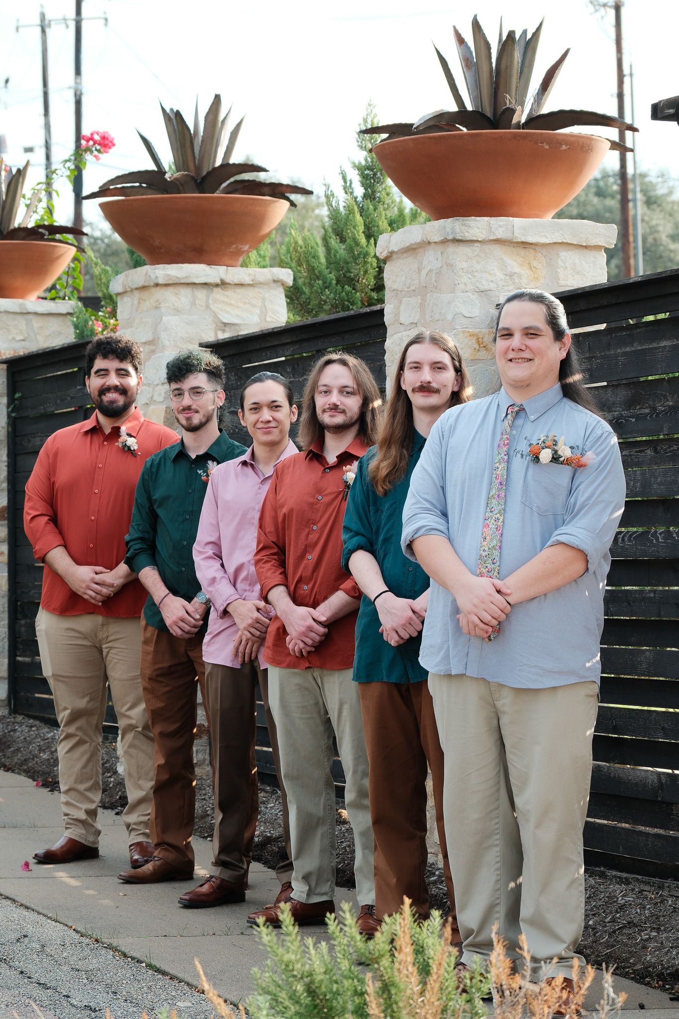 Six groomsmen in colorful button-up shirts pose together outdoors with stone pillars and potted agave plants behind them.