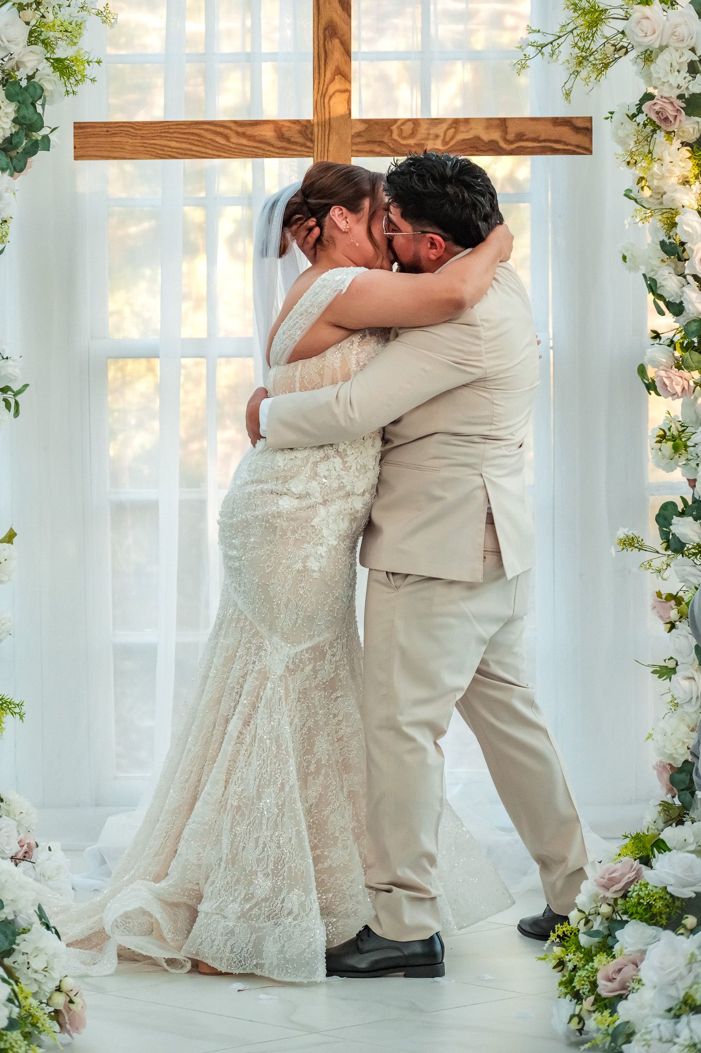 Bride and groom share first kiss embracing under floral arch with wooden cross at wedding ceremony.