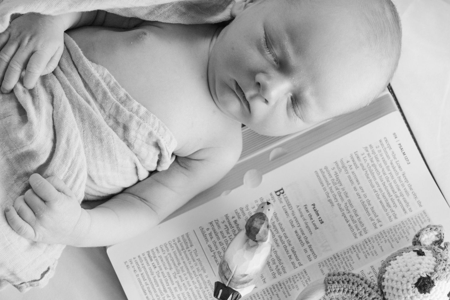 Black and white photo of a sleeping newborn baby lying next to an open book and crocheted toy booties.