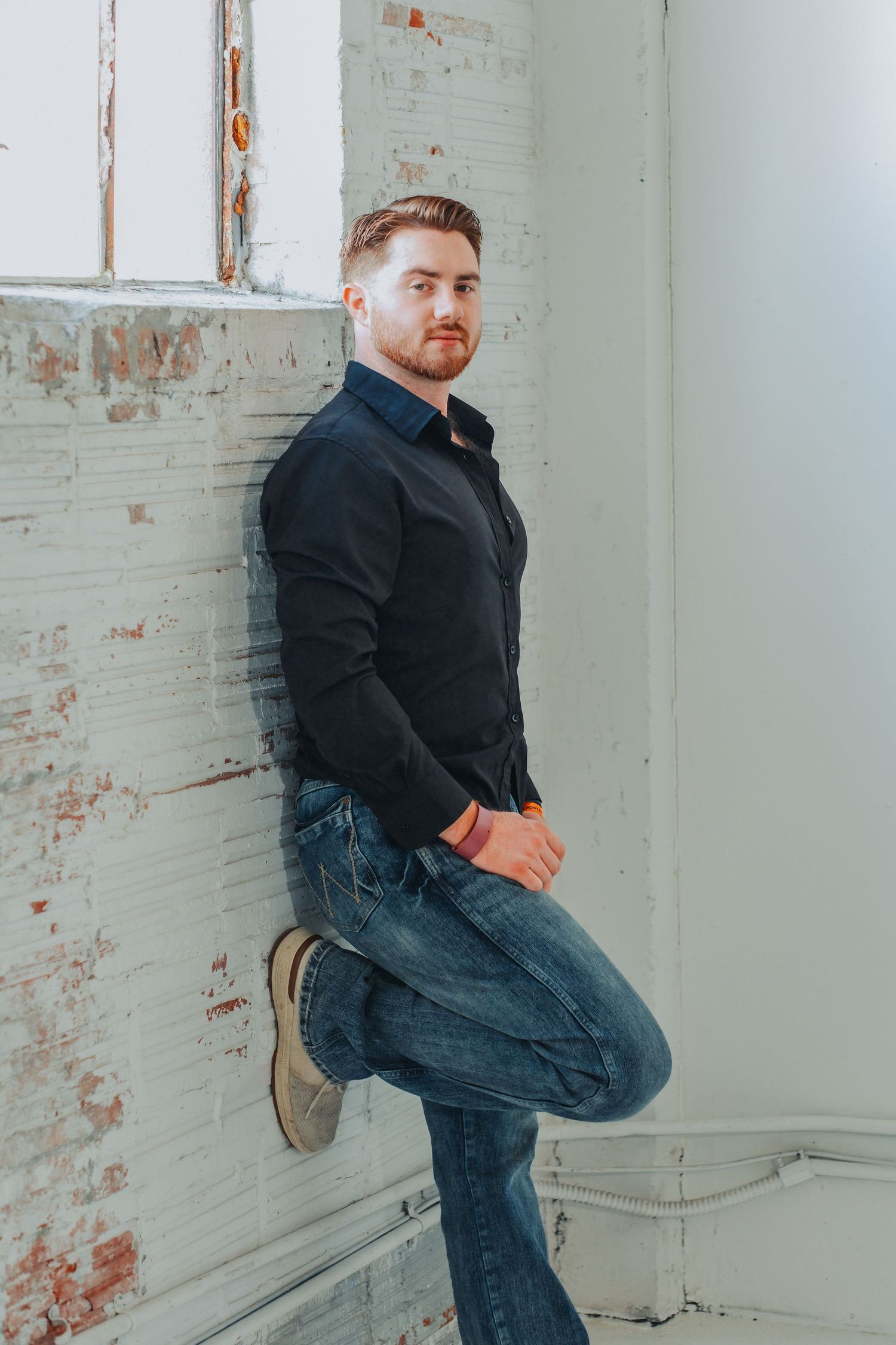 Man in black shirt and blue jeans posing against a white brick wall in a casual portrait.