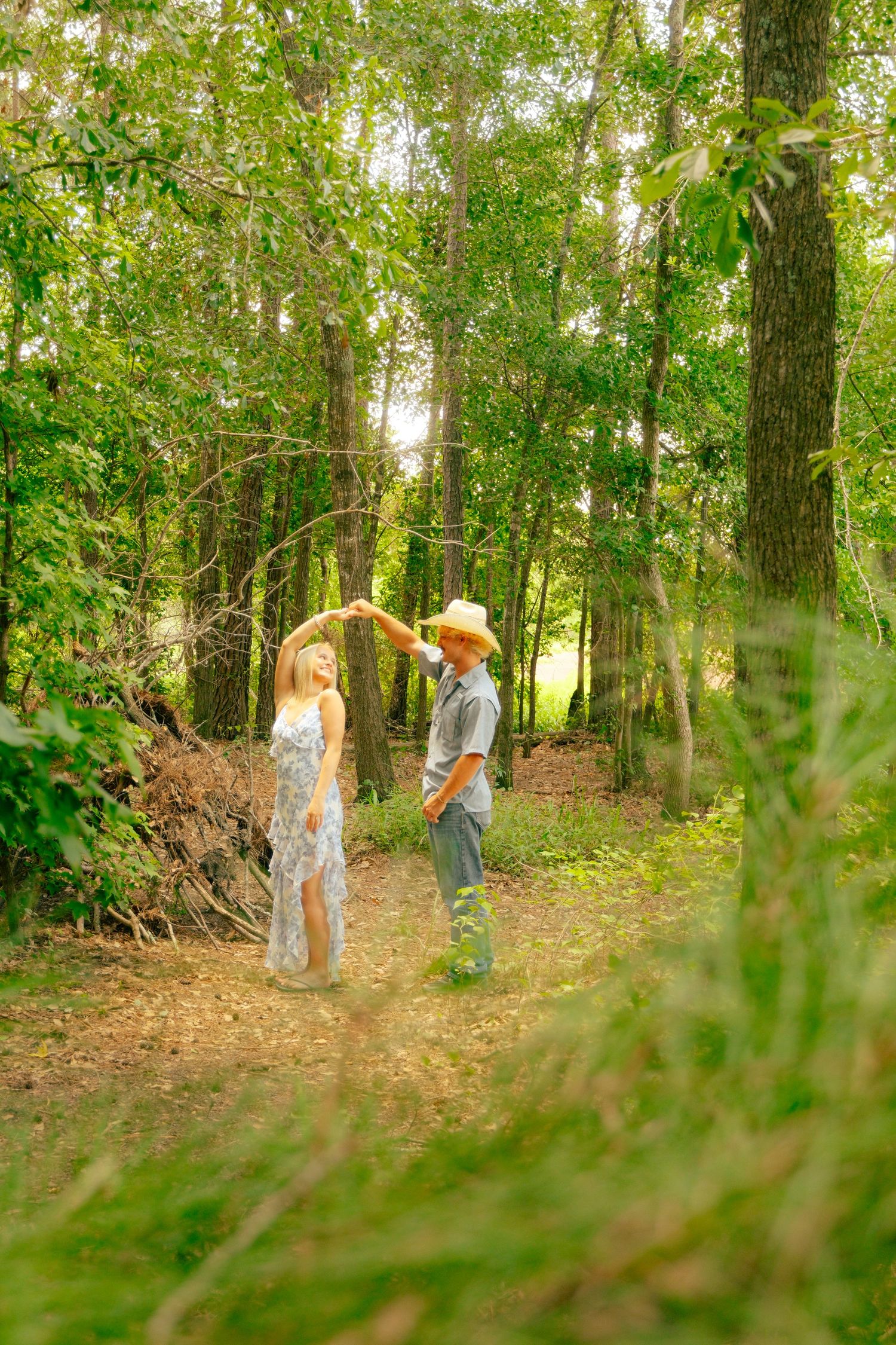 A couple dancing together in a sunlit forest, the woman in a floral dress being twirled by a man in a cowboy hat.