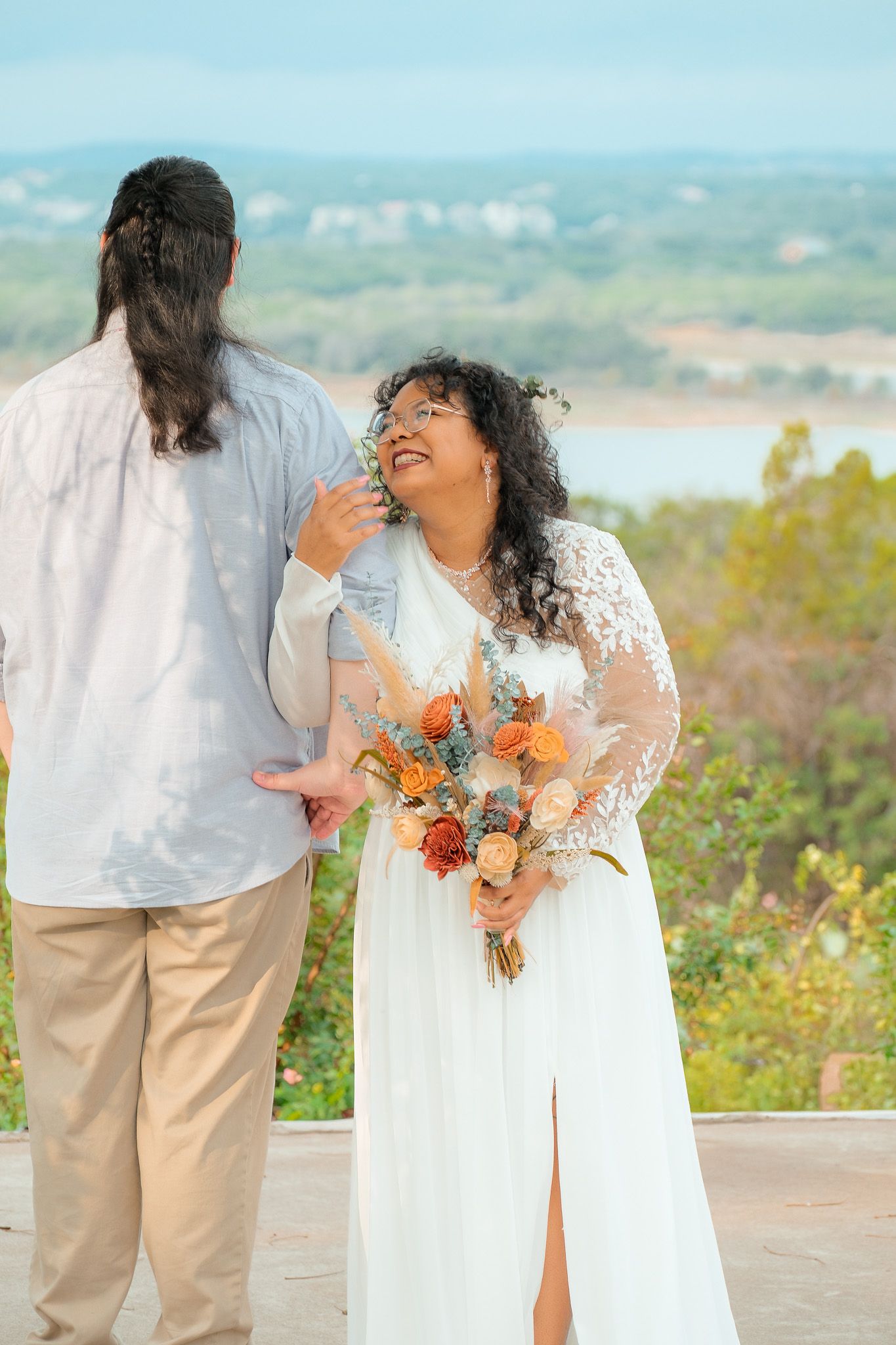 A bride in a lace wedding dress holds an orange and pink bouquet while smiling at her partner outdoors.