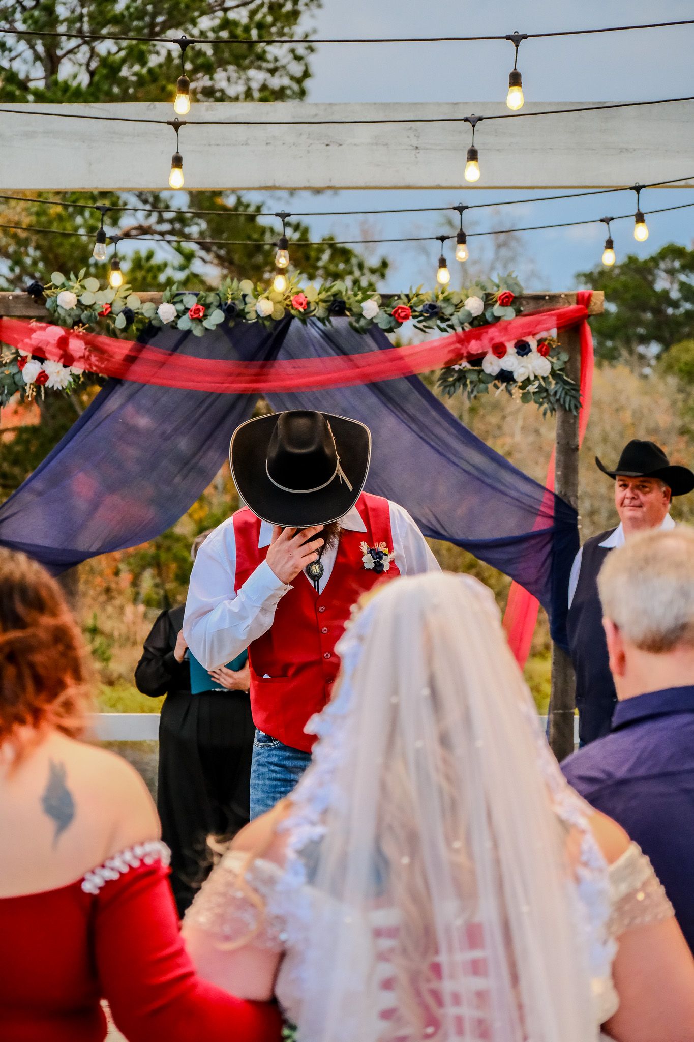 Outdoor western wedding ceremony with groom in cowboy hat and red vest kissing bride under floral arch with string lights.