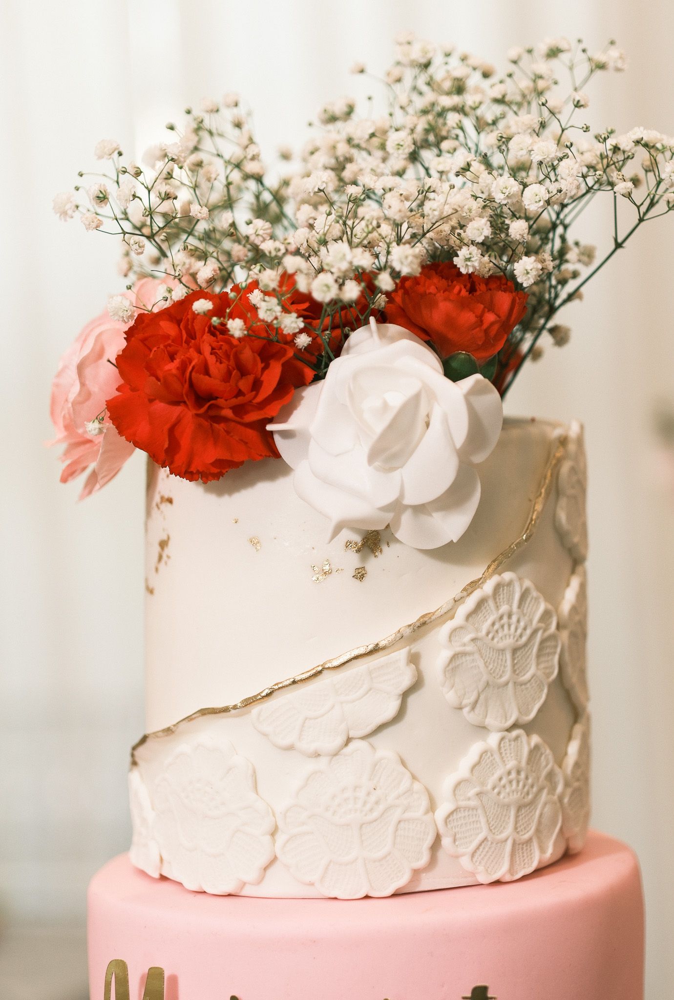 Two-tier wedding cake with white lace texture, gold details, and red carnation and baby's breath topper.