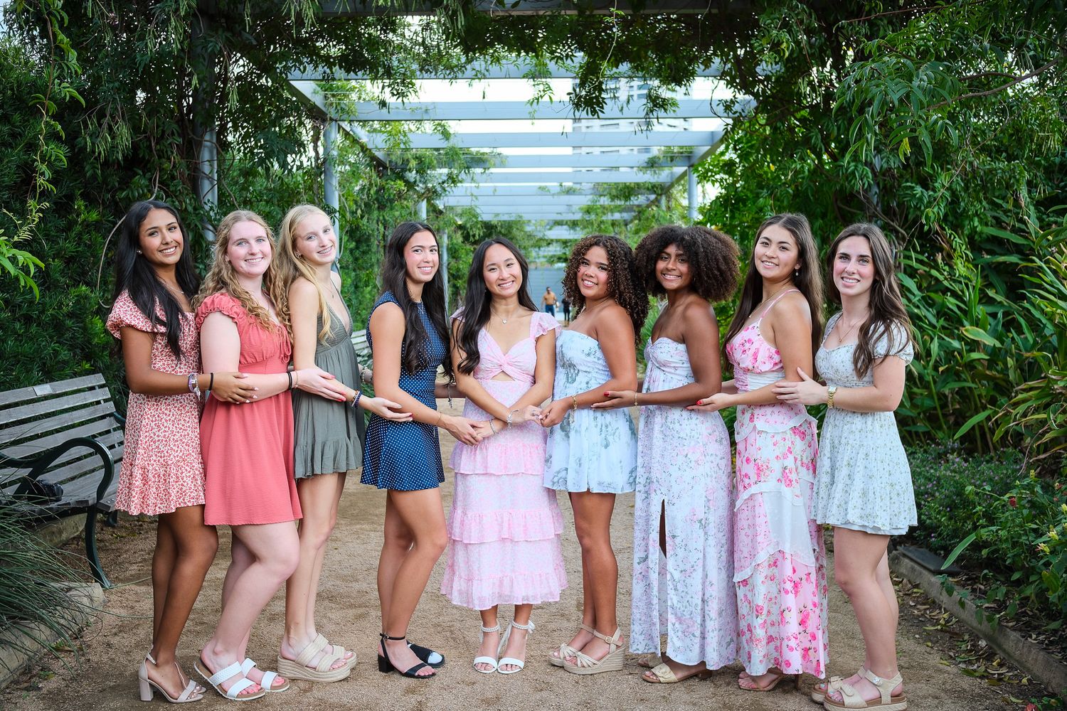 Nine young women in colorful summer dresses posing together under a green garden archway.
