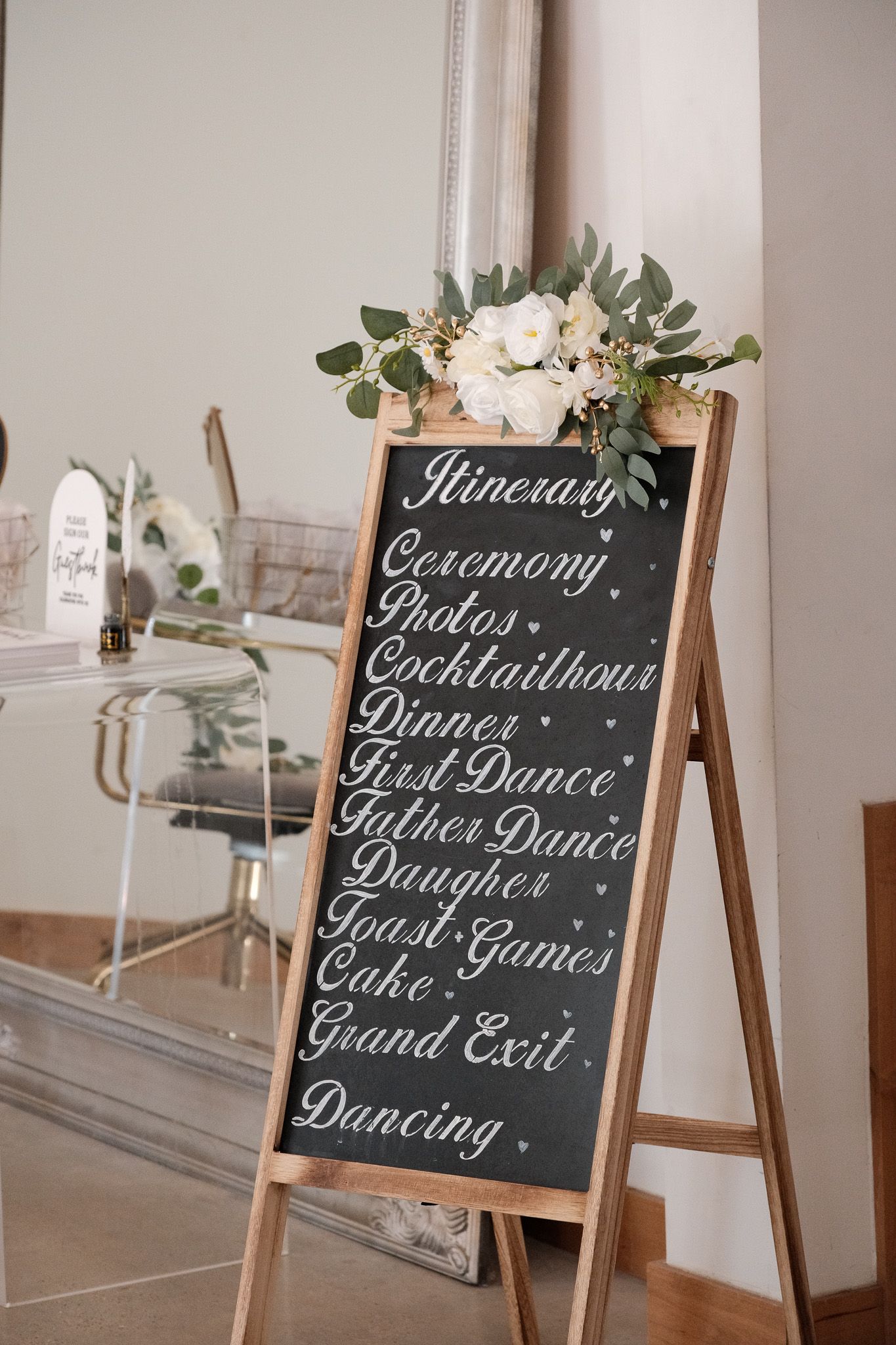 Elegant chalkboard wedding itinerary sign decorated with white flowers and greenery on a wooden easel.