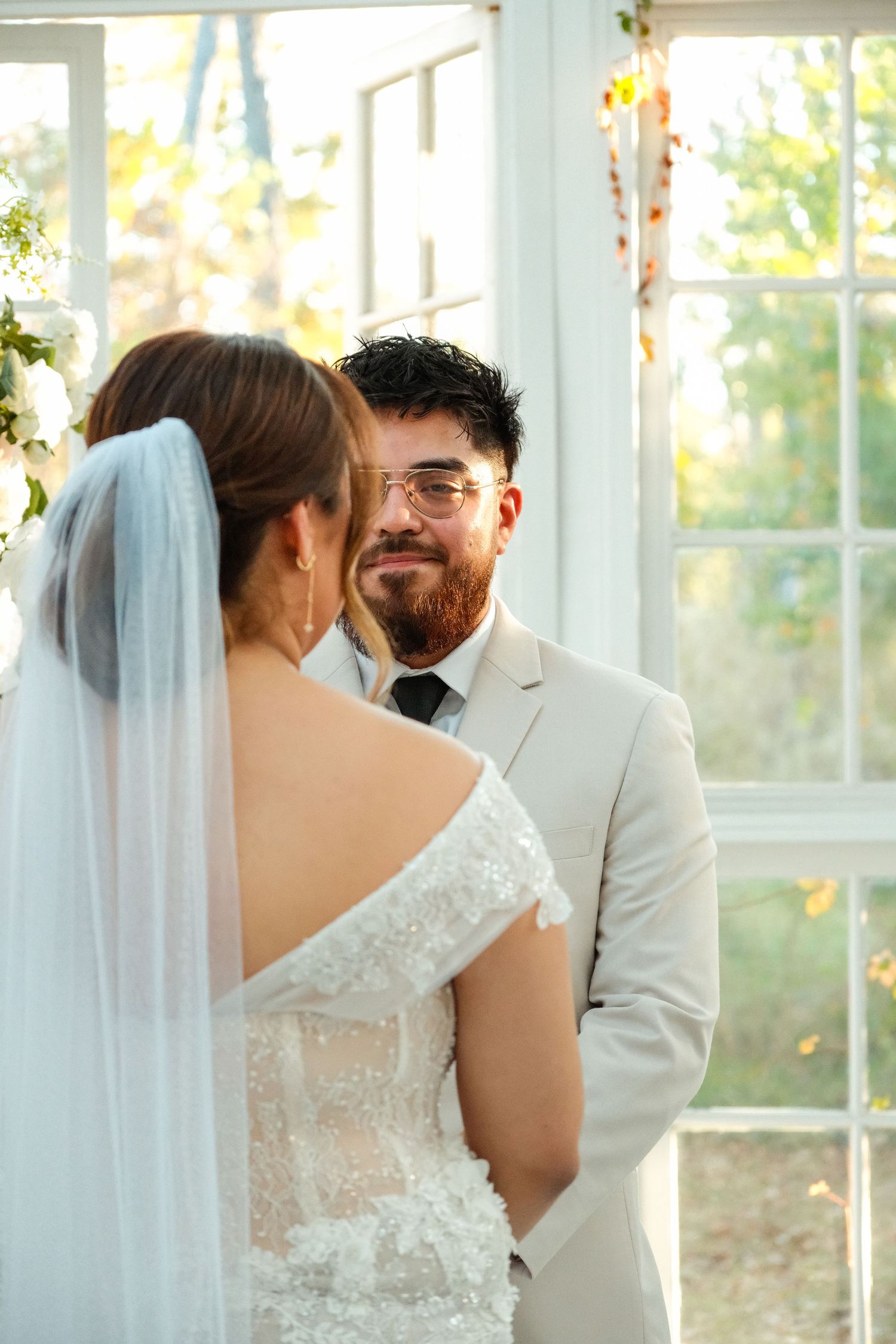 Smiling bearded groom in cream suit gazing at bride during indoor wedding ceremony.
