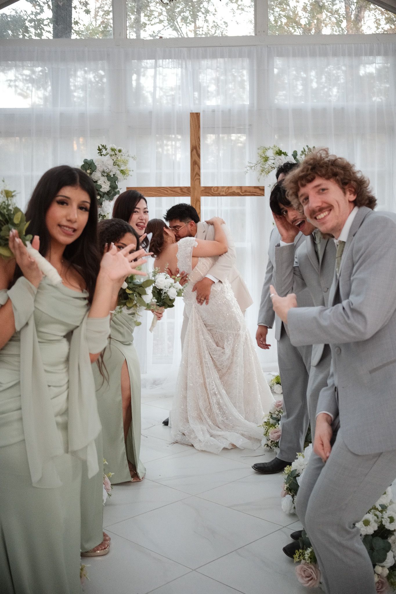 Bridesmaids in sage green dresses and groomsmen pose as bride and groom embrace at wedding altar.