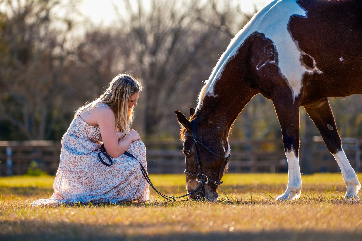 Pregnant woman in floral dress sitting beside a paint horse grazing in golden light.