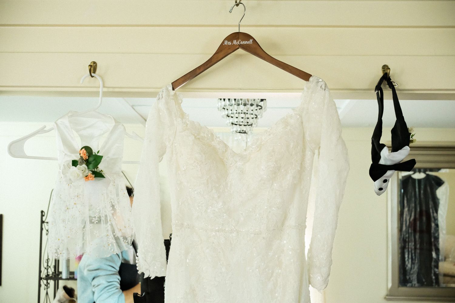 White lace long-sleeve wedding dress hanging on wooden hanger in bridal suite with accessories nearby.