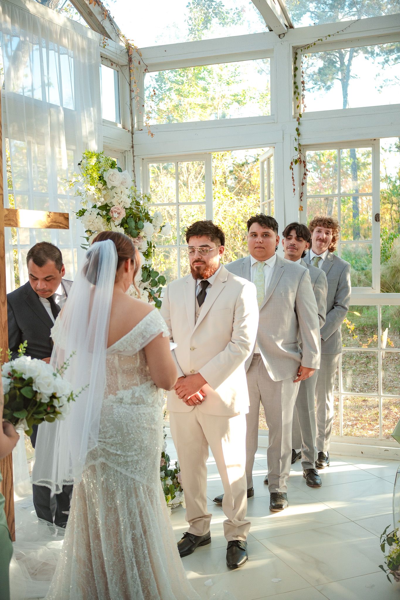 Groom in cream suit watching bride approach altar in sunlit greenhouse wedding ceremony venue.