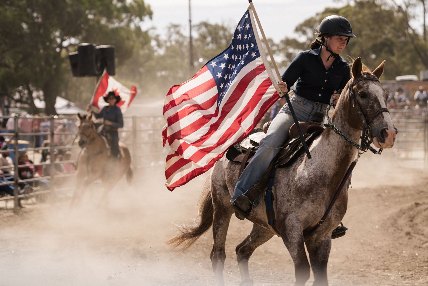 A rider on horseback carries an American flag through a dusty rodeo arena.